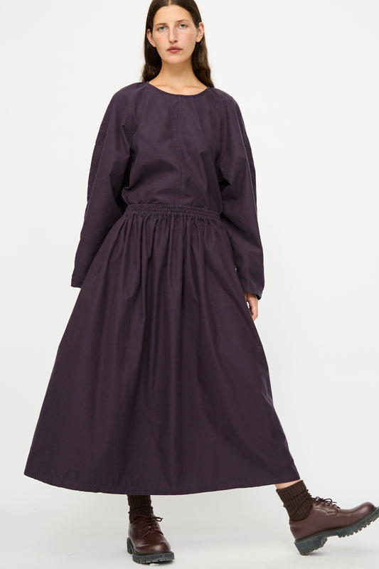 A person wears the SONO Skye Skirt in Dark Grape, styled with a loose dark purple dress, brown socks, and brown lace-up shoes, standing against a plain white background.