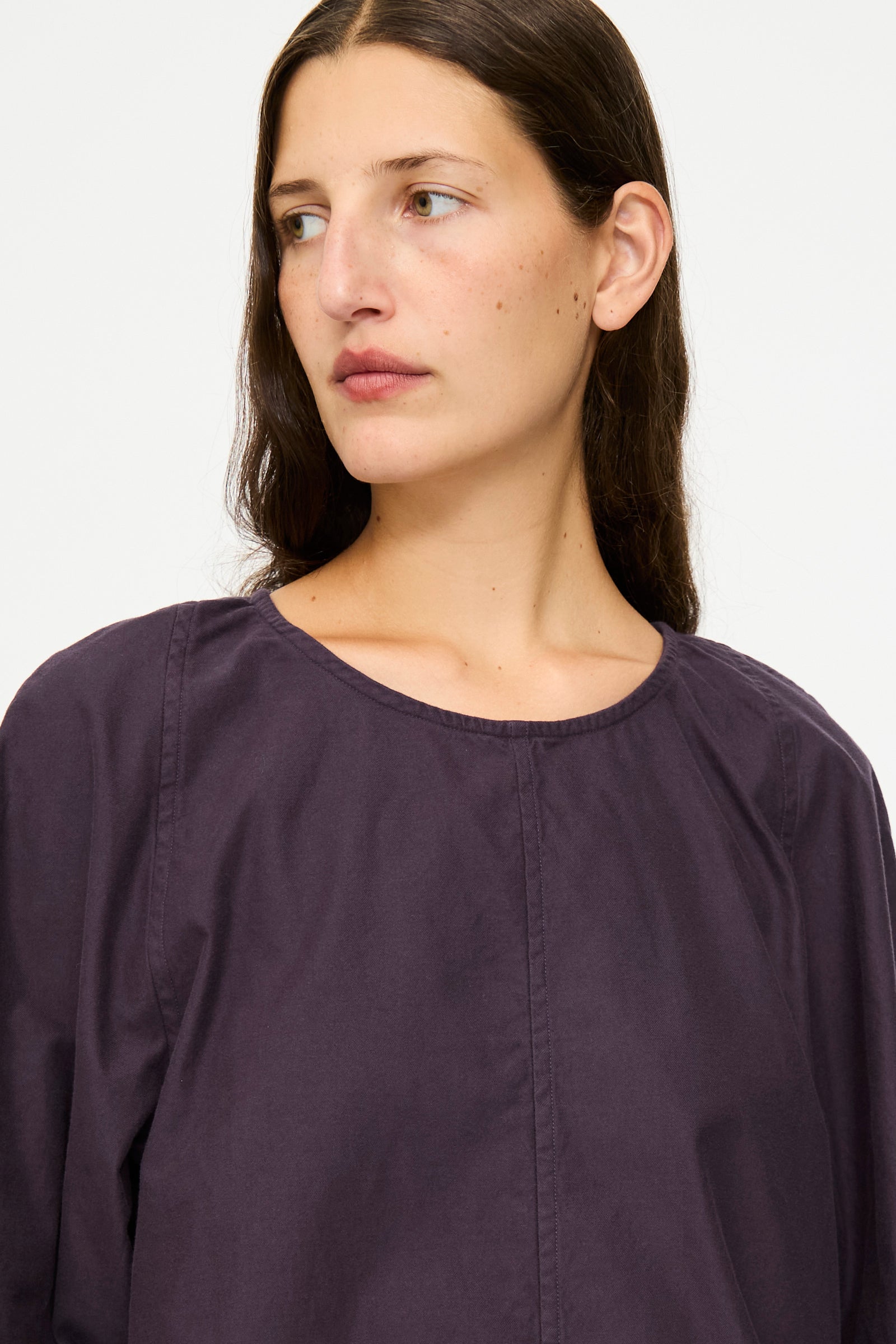 Woman with long brown hair wears the SONO Tiga Top in Dark Grape, an oversized cotton poplin shirt, while looking slightly to the side against a plain background.