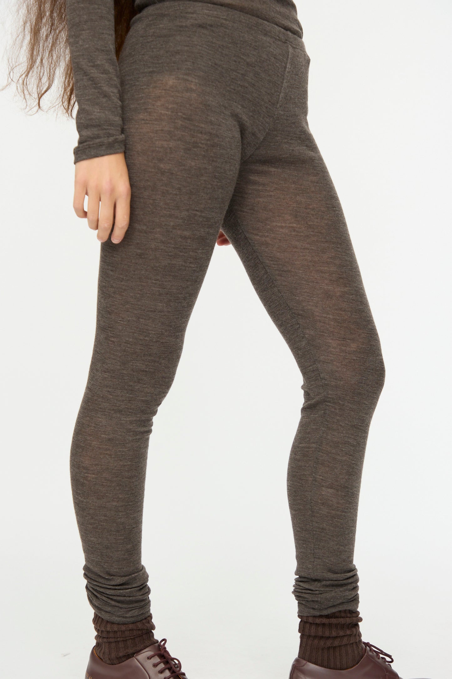 A person wears SONO’s Wool Lupo Leggings in Charcoal with a matching long sleeve top, brown socks, and dark brown shoes, standing against a plain white background.