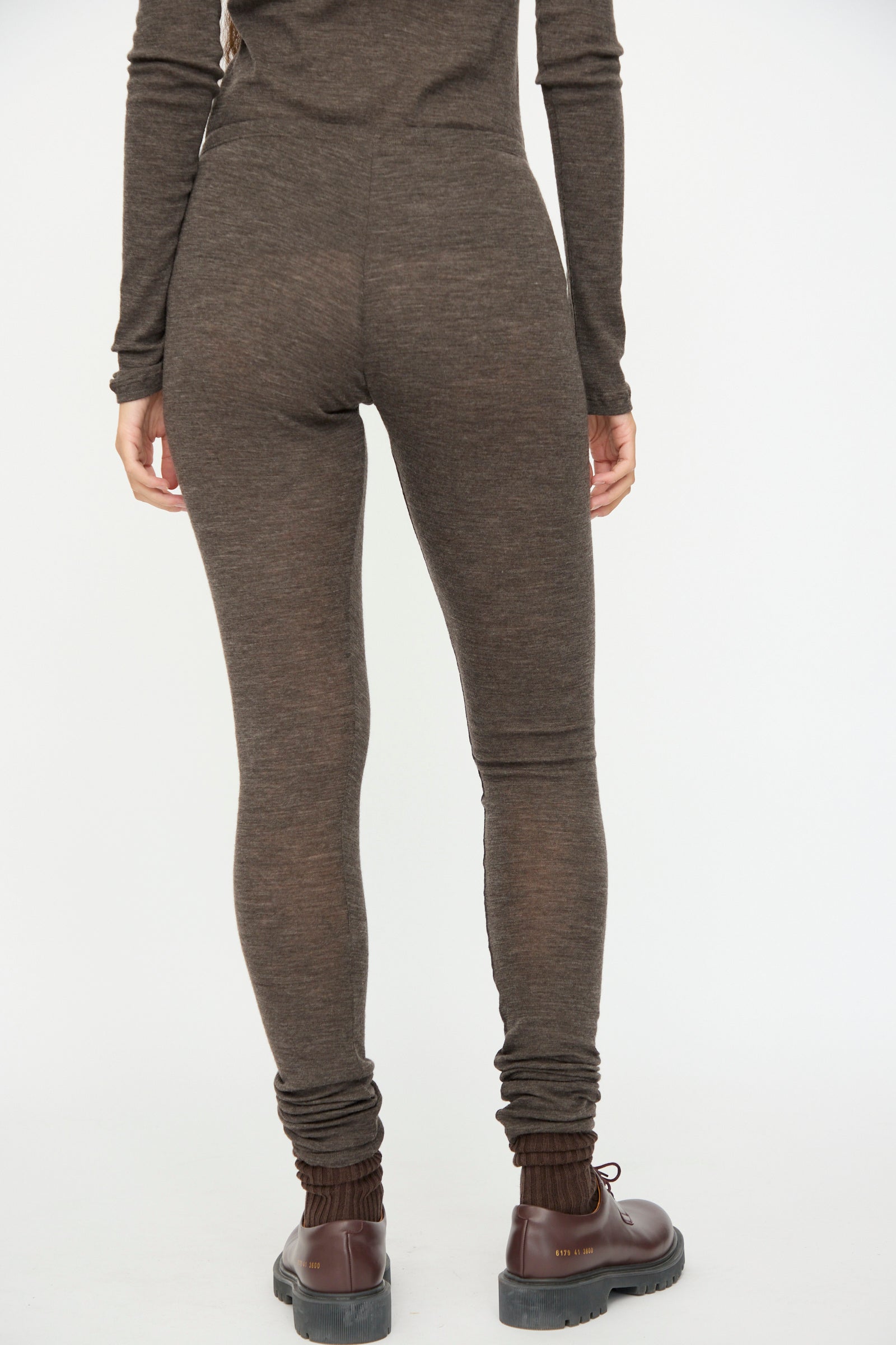 A person faces away from the camera, wearing SONO's Wool Lupo Leggings in Charcoal, a fitted ribbed style made in Italy, paired with a long-sleeve top and brown ankle boots against a plain white background.