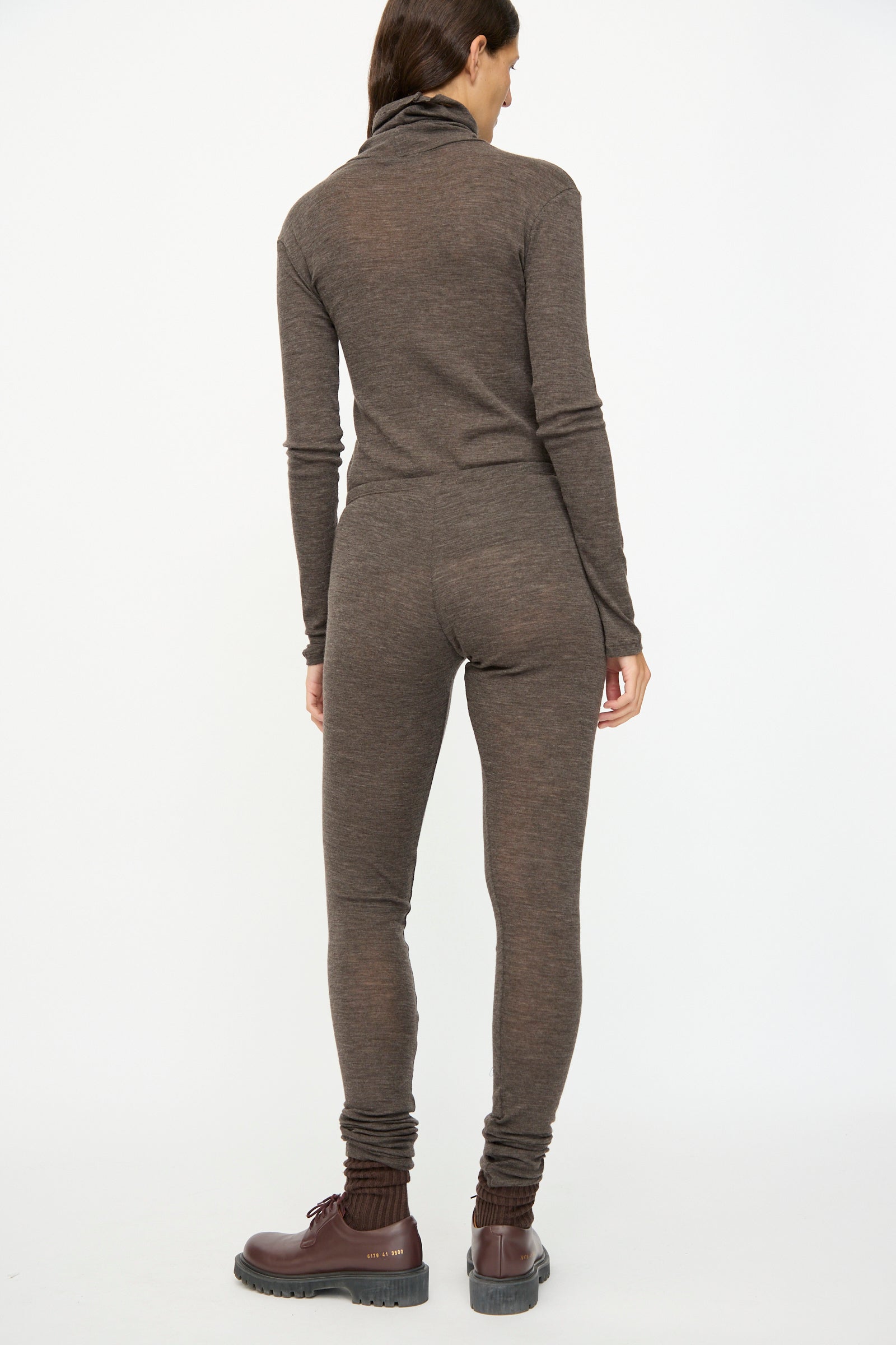 A person stands facing away from the camera against a white background, wearing SONO Wool Lupo Leggings in Charcoal, styled with brown long-sleeve fitted jumpsuit and brown lace-up boots; ribbed details add texture.