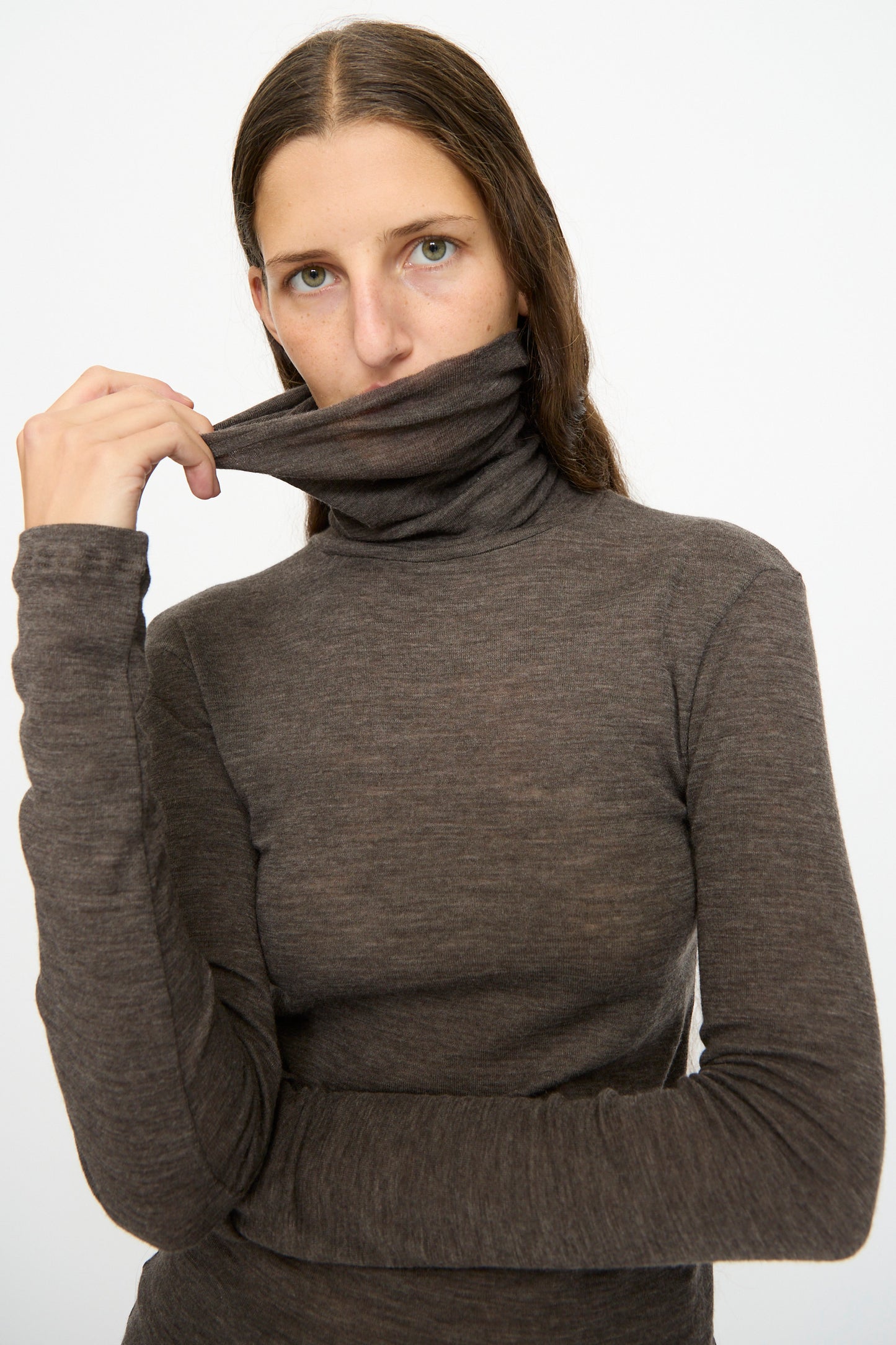 A person with long brown hair, wearing the SONO Wool Roberta Rollneck in Charcoal and pulling the collar up to partially cover their mouth, stands against a plain white background.