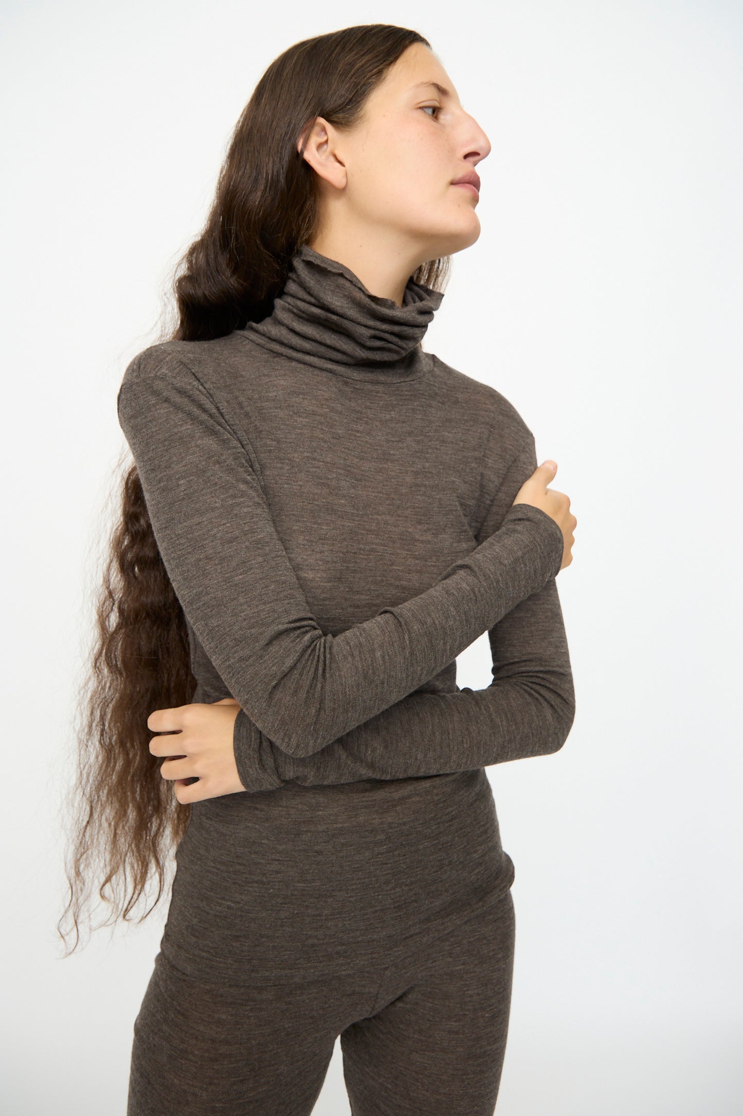 Model wearing the SONO Wool Roberta Rollneck in Charcoal, featuring long sleeves, paired with slim fit pants, standing against a plain light background with long hair draped over one shoulder.
