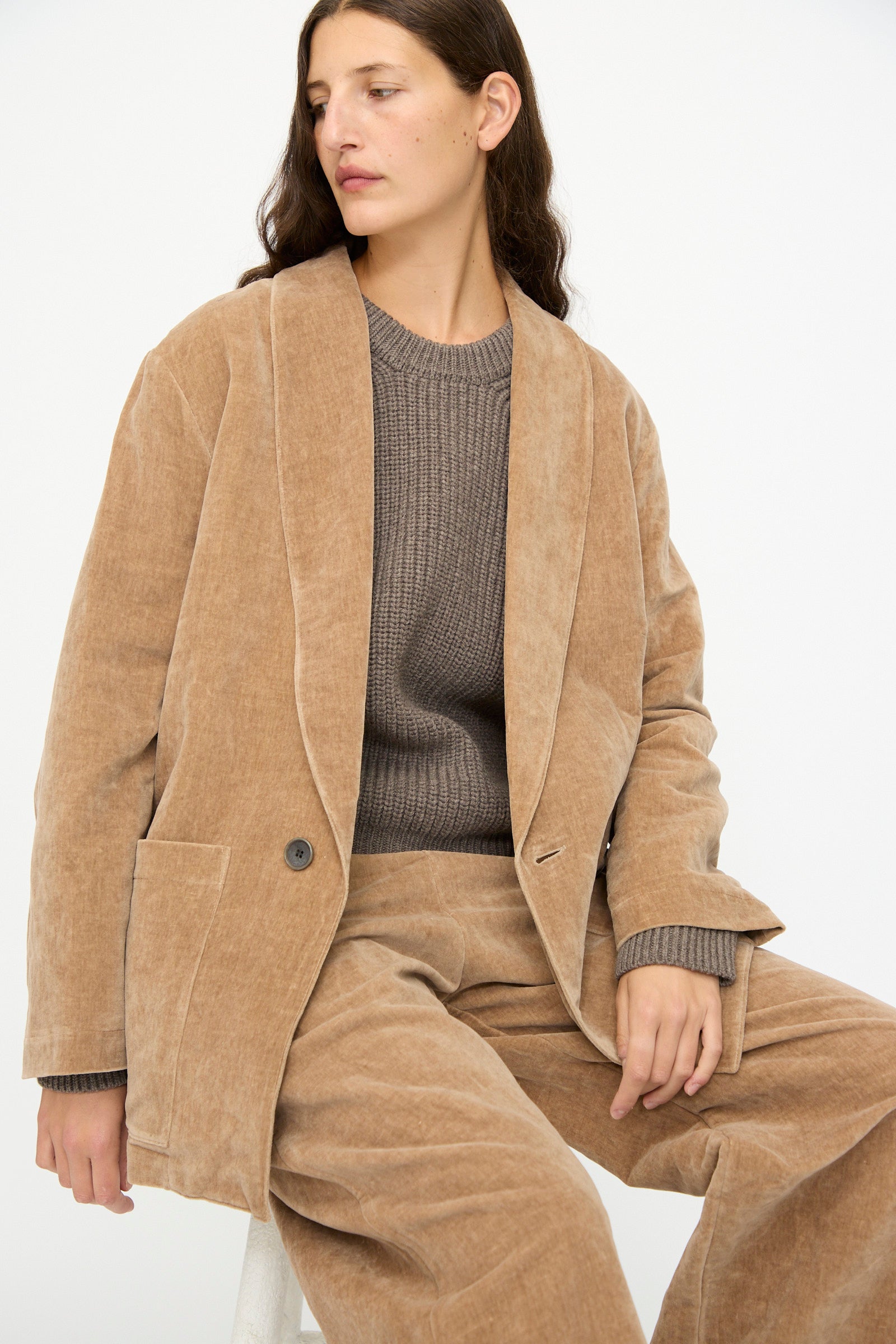 A woman wears the SONO Yoyogi Jacket in Desert Rose, seated on a white stool against a plain background.