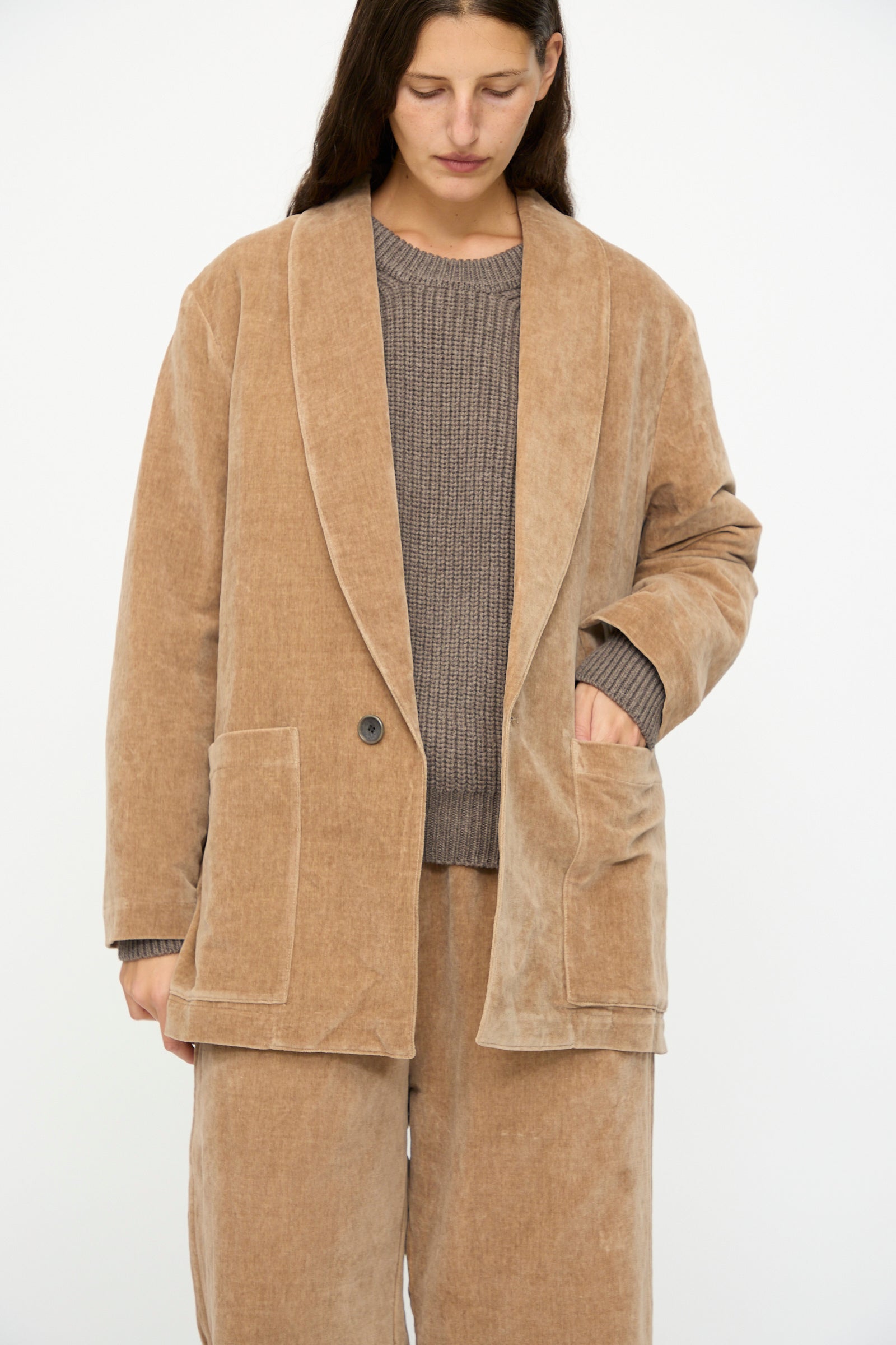 A person wearing SONO's Yoyogi Jacket in Desert Rose styled with matching pants over a textured brown sweater, standing against a plain white background.