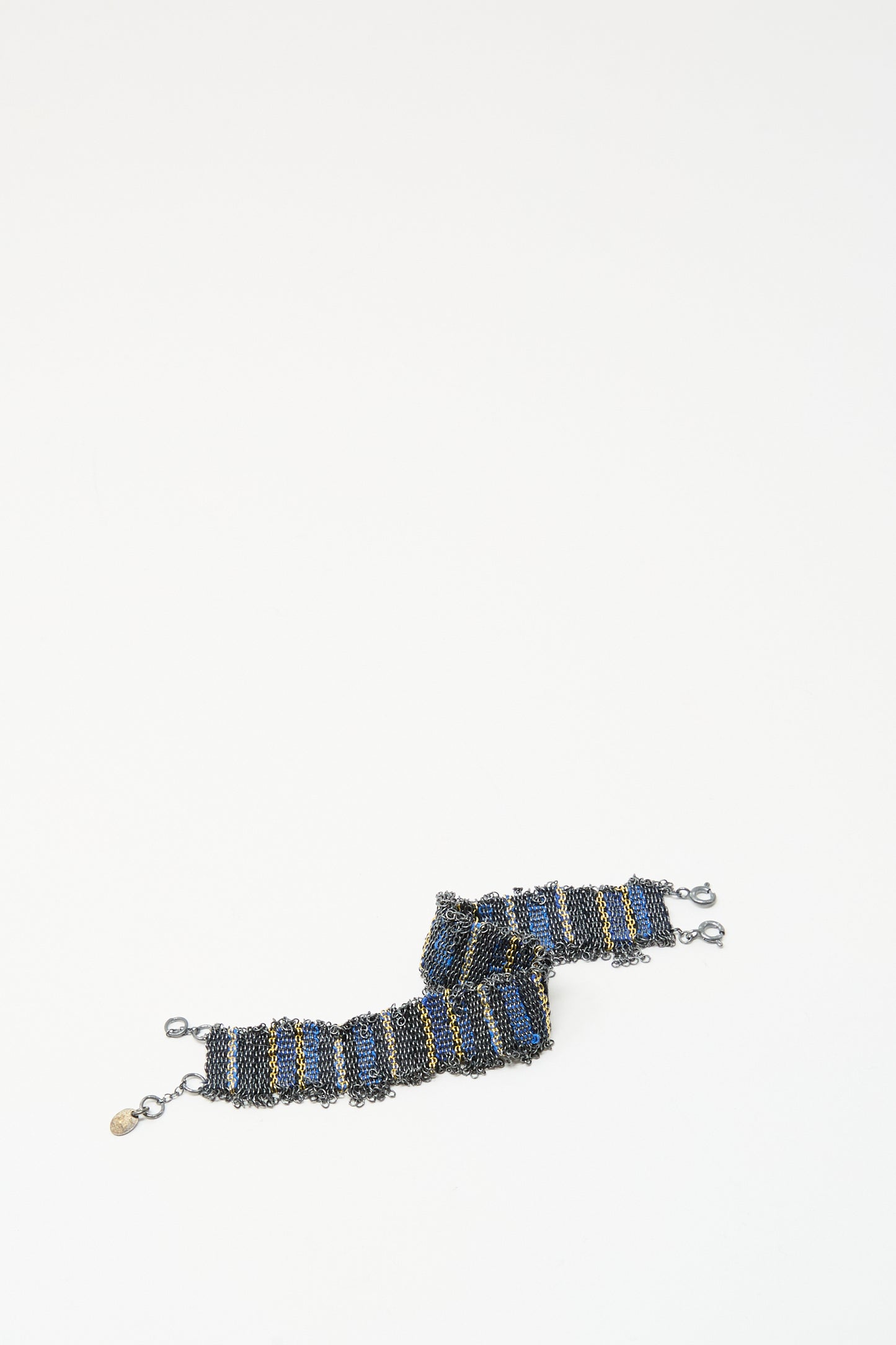 The Stephanie Schneider Silver and Silk Woven Bracelet in Blue features blue, black, and gold stripes with metal clasps and a small gold tag, displayed on a white background.