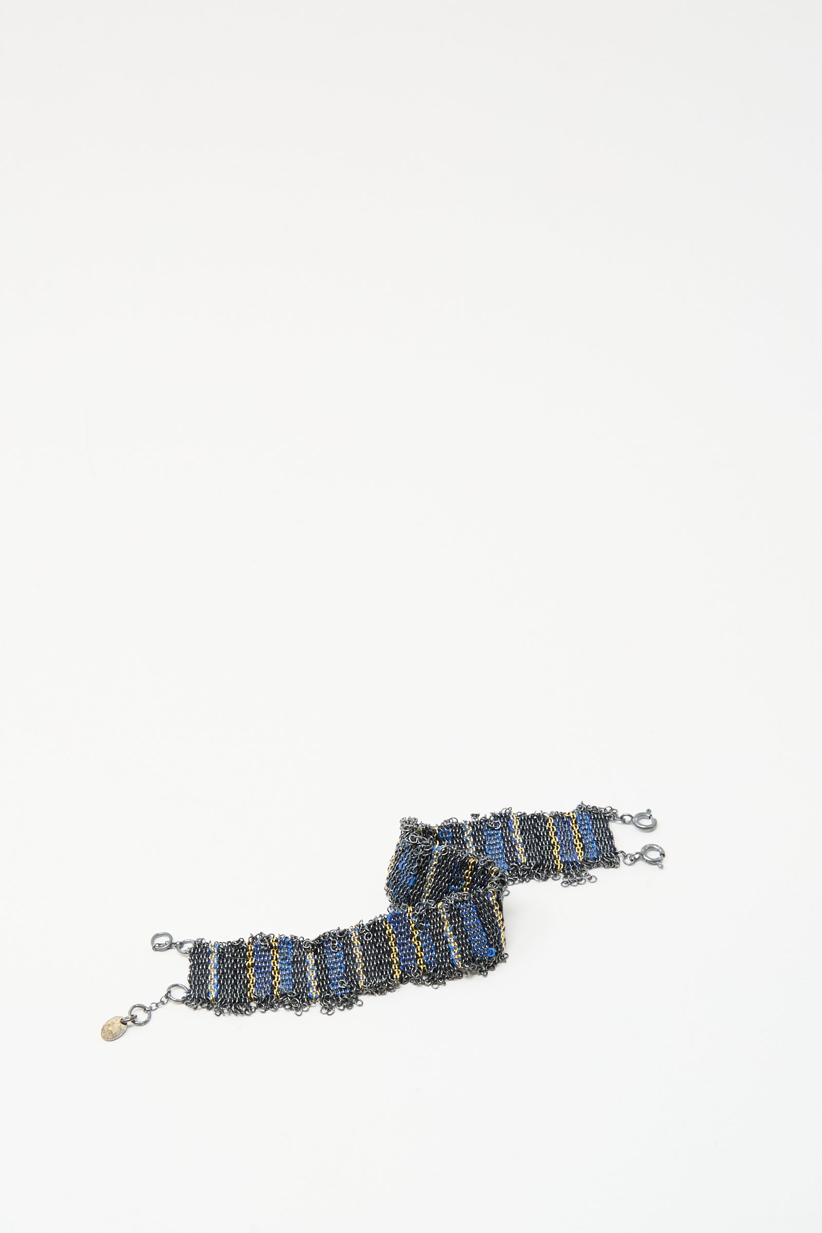 The Stephanie Schneider Silver and Silk Woven Bracelet in Blue features blue, black, and gold stripes with metal clasps and a small gold tag, displayed on a white background.