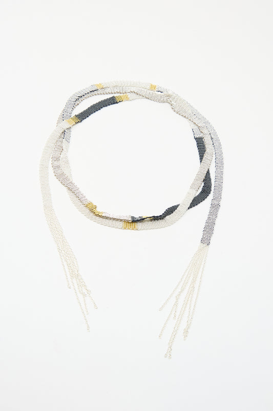 The Silver and Silk Woven Shawl Necklace by Stephanie Schneider features alternating white, gold, and black beads with loose strands on both ends, reminiscent of a sterling silver shawl, displayed on a white background.