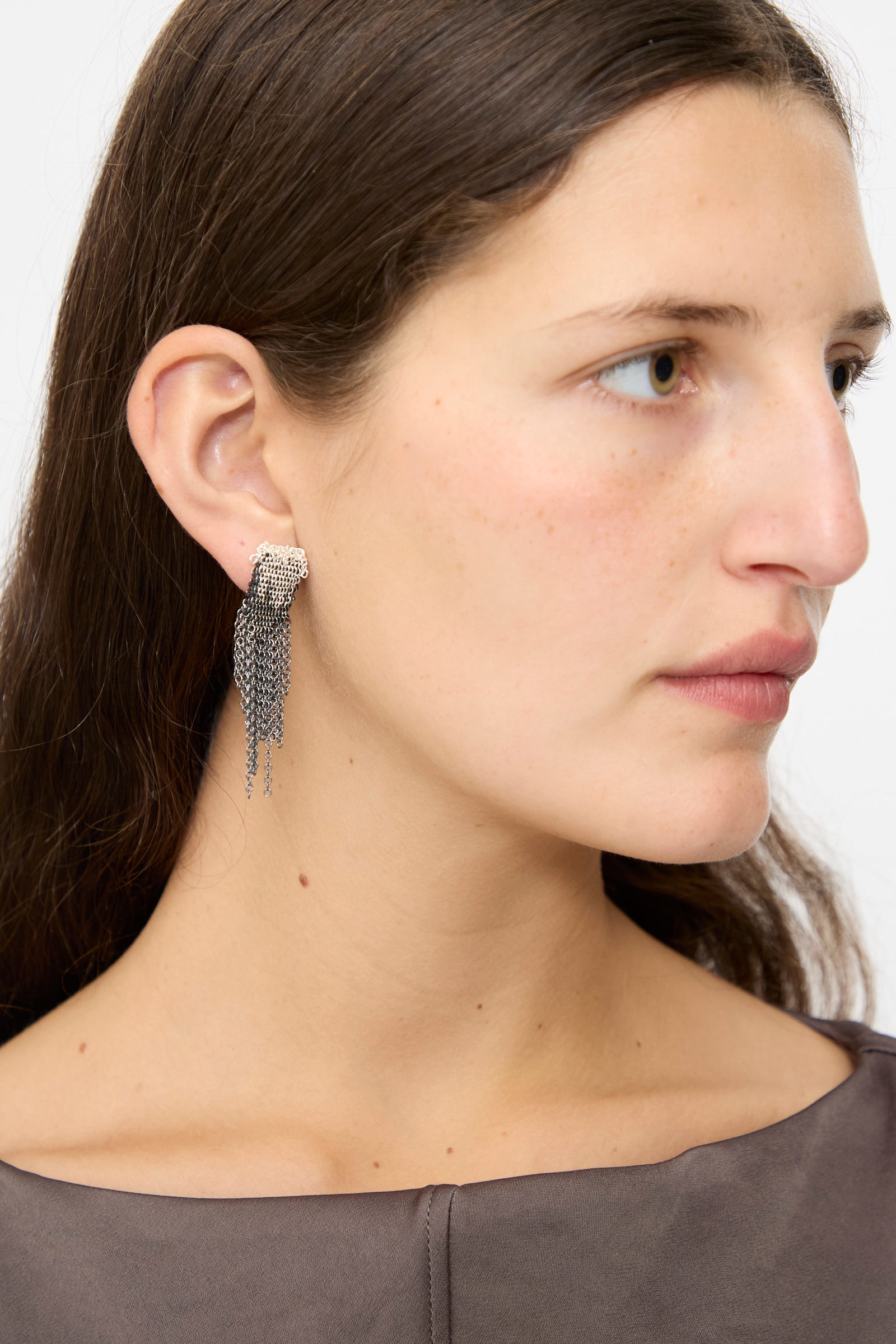 A woman with long brown hair wears Stephanie Schneider's Sterling Silver Oxidized and Silk Earrings in Anthracite, featuring multiple dangling strands, shown in a close-up side profile.