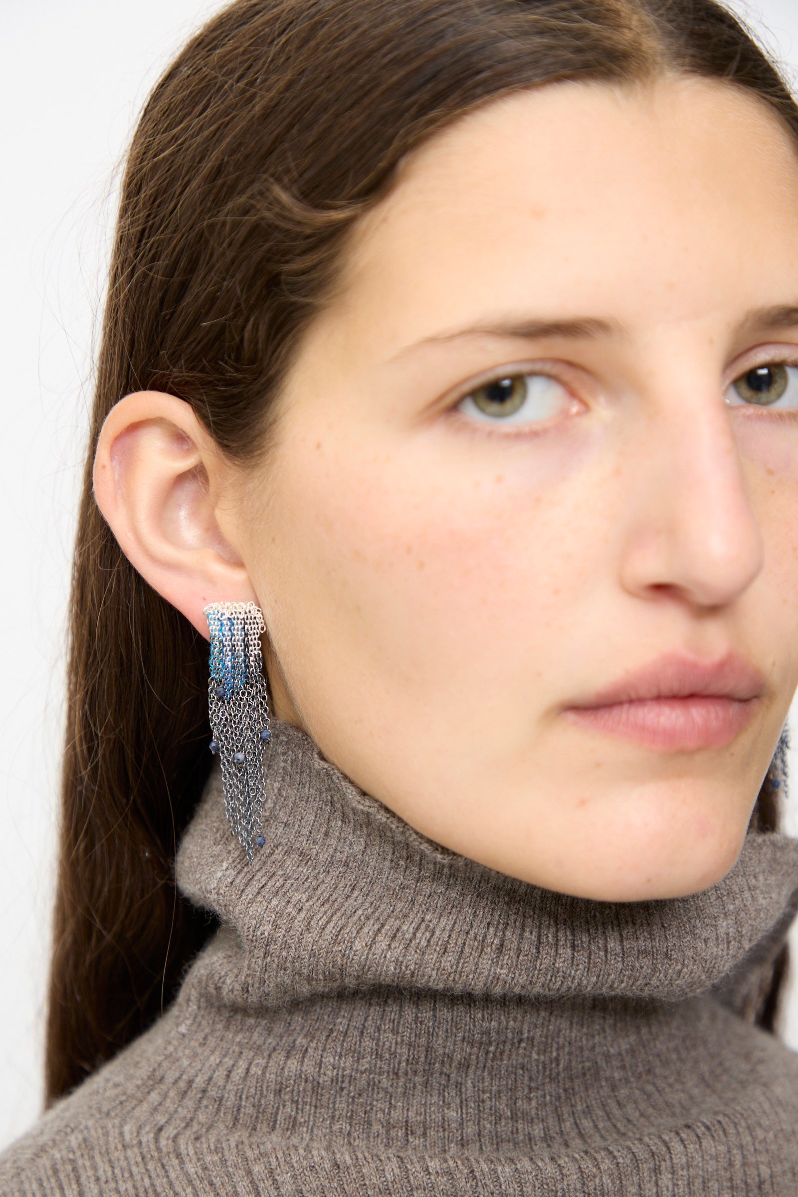 A woman with straight brown hair wears a taupe turtleneck and Stephanie Schneider Sterling Silver and Silk Earrings in Blue with Sapphires, looking toward the camera.