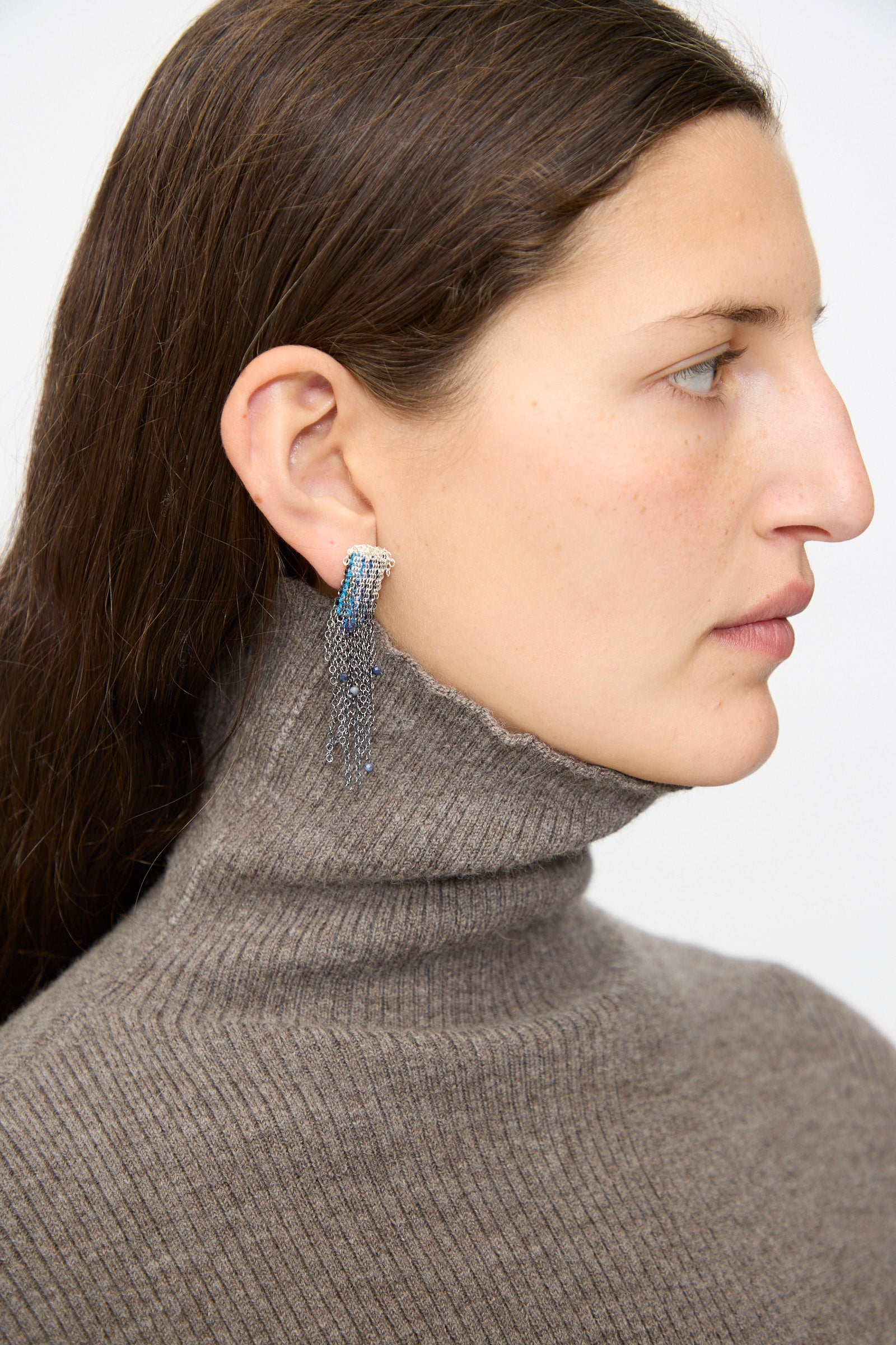 A woman with long brown hair wears a grey turtleneck and models the Sterling Silver and Silk Earrings in Blue with Sapphires by Stephanie Schneider, shown in profile against a plain background.