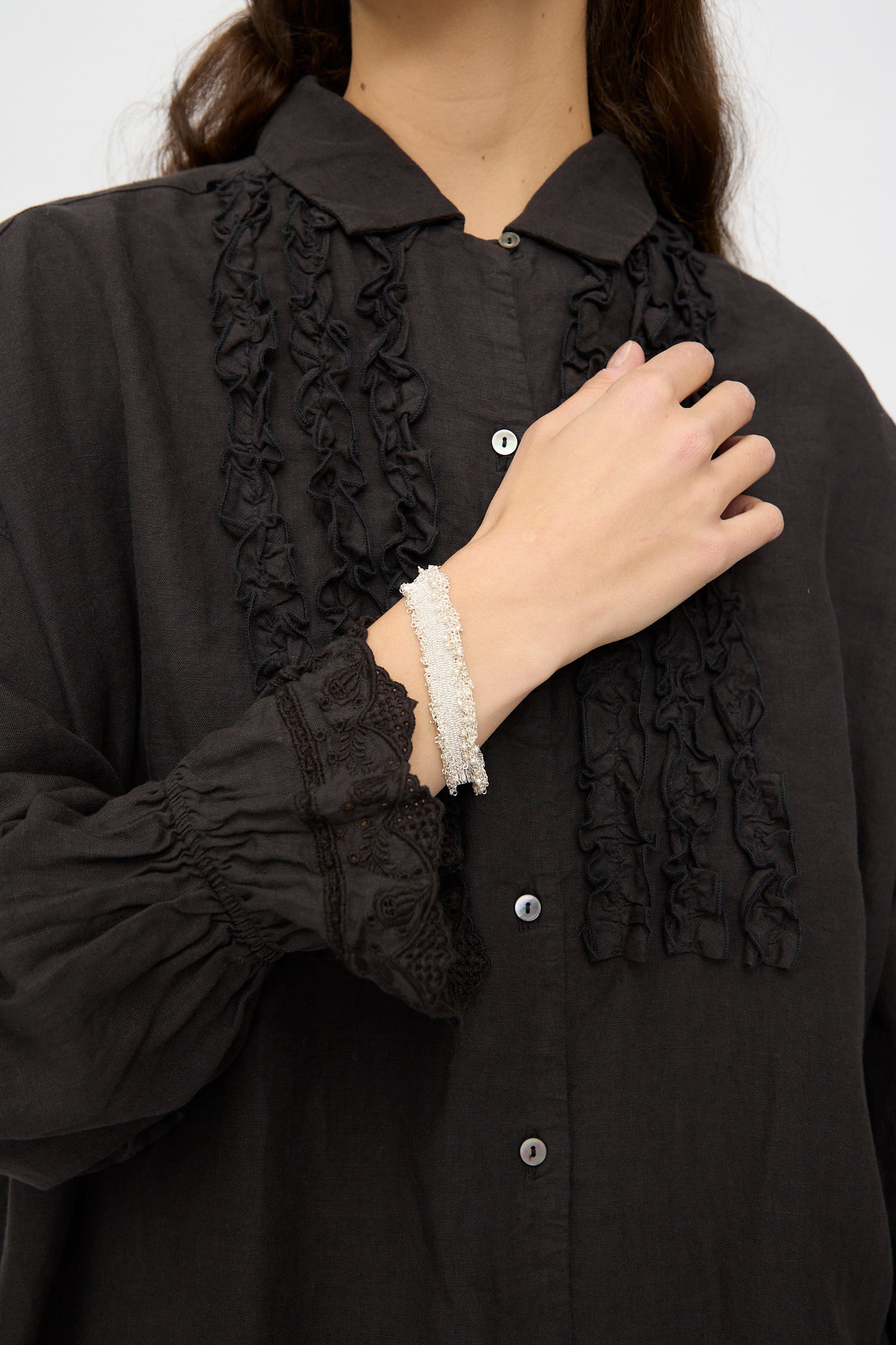 A person wearing a black, ruffled, button-up shirt with lace details displays the Stephanie Schneider Silver and Silk Woven Bracelet with Pearls on their wrist.