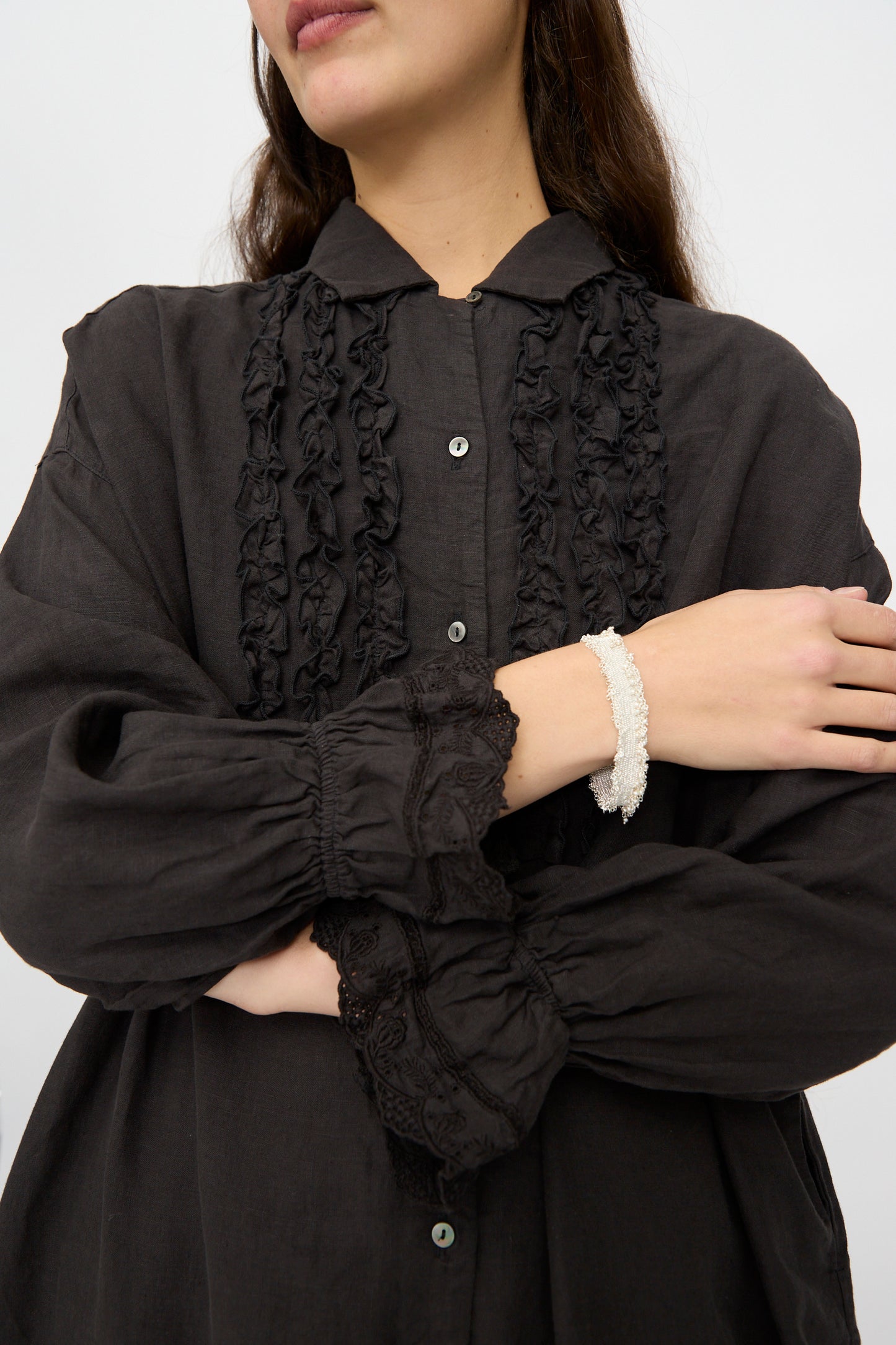 A woman crosses her arms, highlighting the Stephanie Schneider Silver and Silk Woven Bracelet with Pearls on her wrist as she wears a black blouse with ruffled details and lace cuffs.