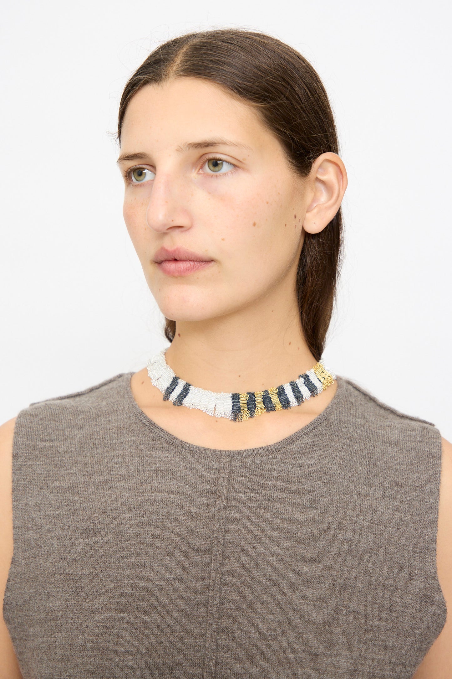 A woman with straight brown hair models Stephanie Schneider’s Silver, Gold and Silk Woven Necklace—a gold-plated silver choker featuring white, black, gold, and gray stripes—paired with a sleeveless taupe top against a plain background.