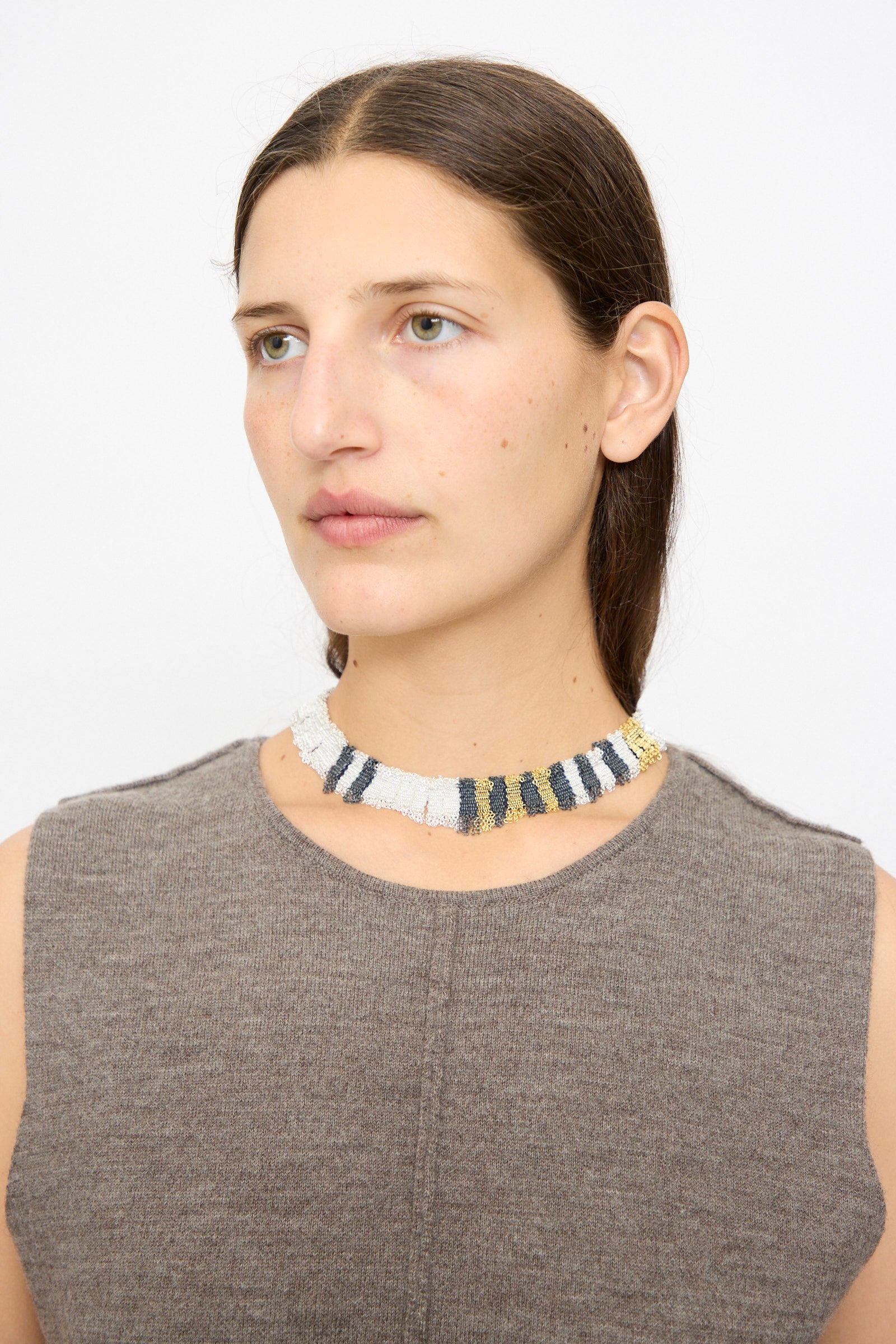 A woman with straight brown hair models Stephanie Schneider’s Silver, Gold and Silk Woven Necklace—a gold-plated silver choker featuring white, black, gold, and gray stripes—paired with a sleeveless taupe top against a plain background.