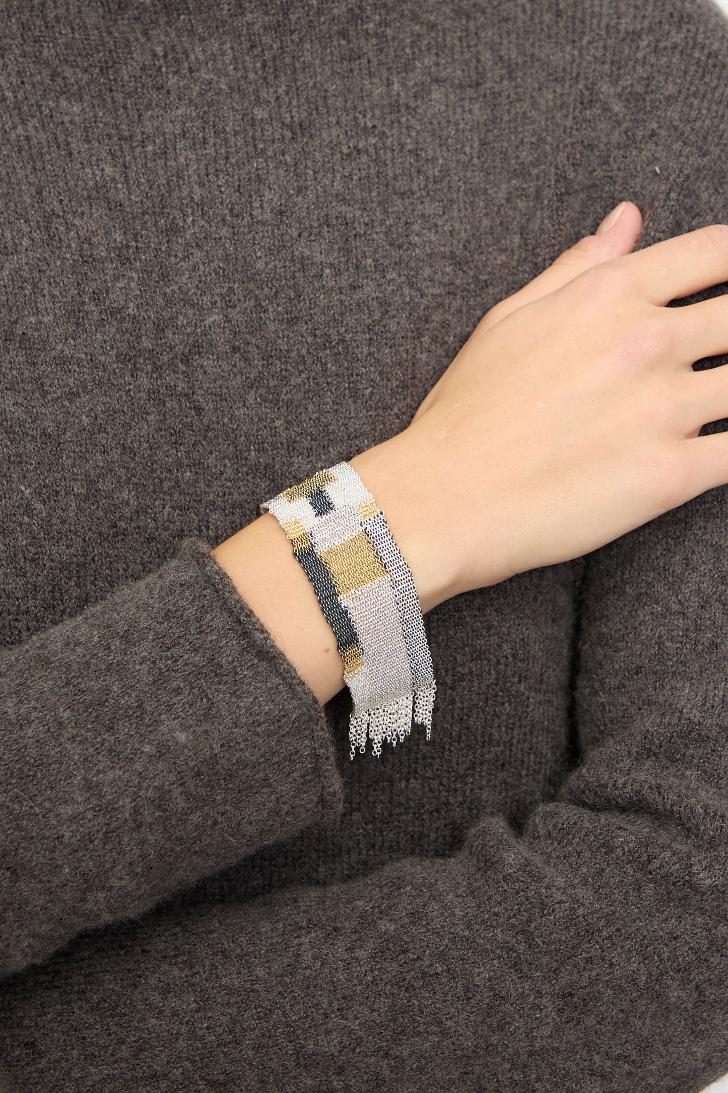 Someone wears the Stephanie Schneider Silver Oxidized, Gold Plated and Silk Woven Bracelet on their wrist over a textured gray long-sleeve top.