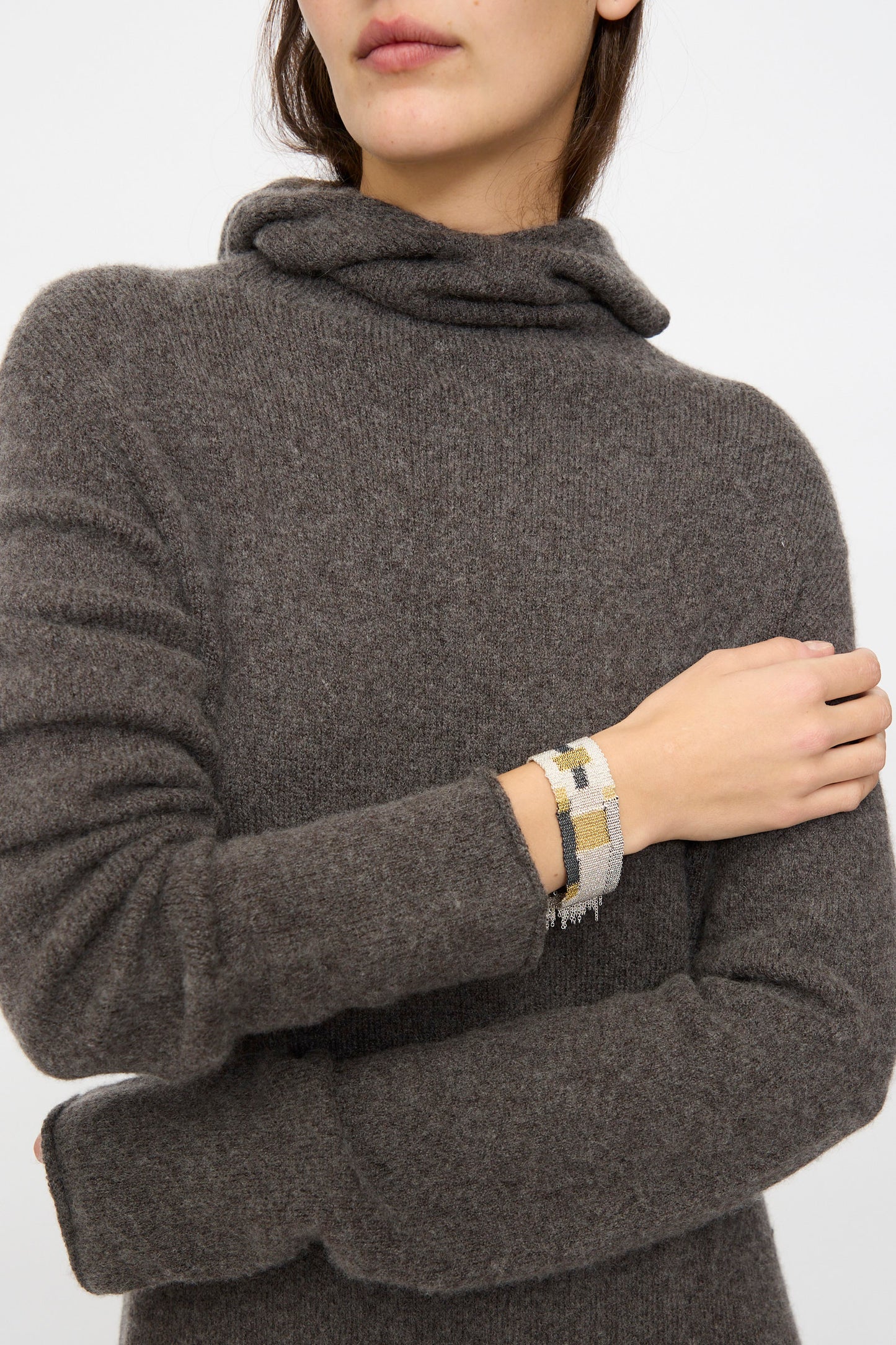 A woman in a brown turtleneck crosses her arms, showcasing a Stephanie Schneider Silver Oxidized, Gold Plated and Silk Woven Bracelet on her wrist.