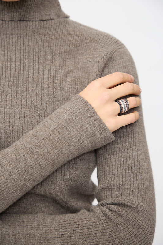 A person wearing a ribbed taupe turtleneck rests their hand on their shoulder, showcasing stacked rings including the Stephanie Schneider Silver, Rose Gold Plated and Silk Woven Ring.