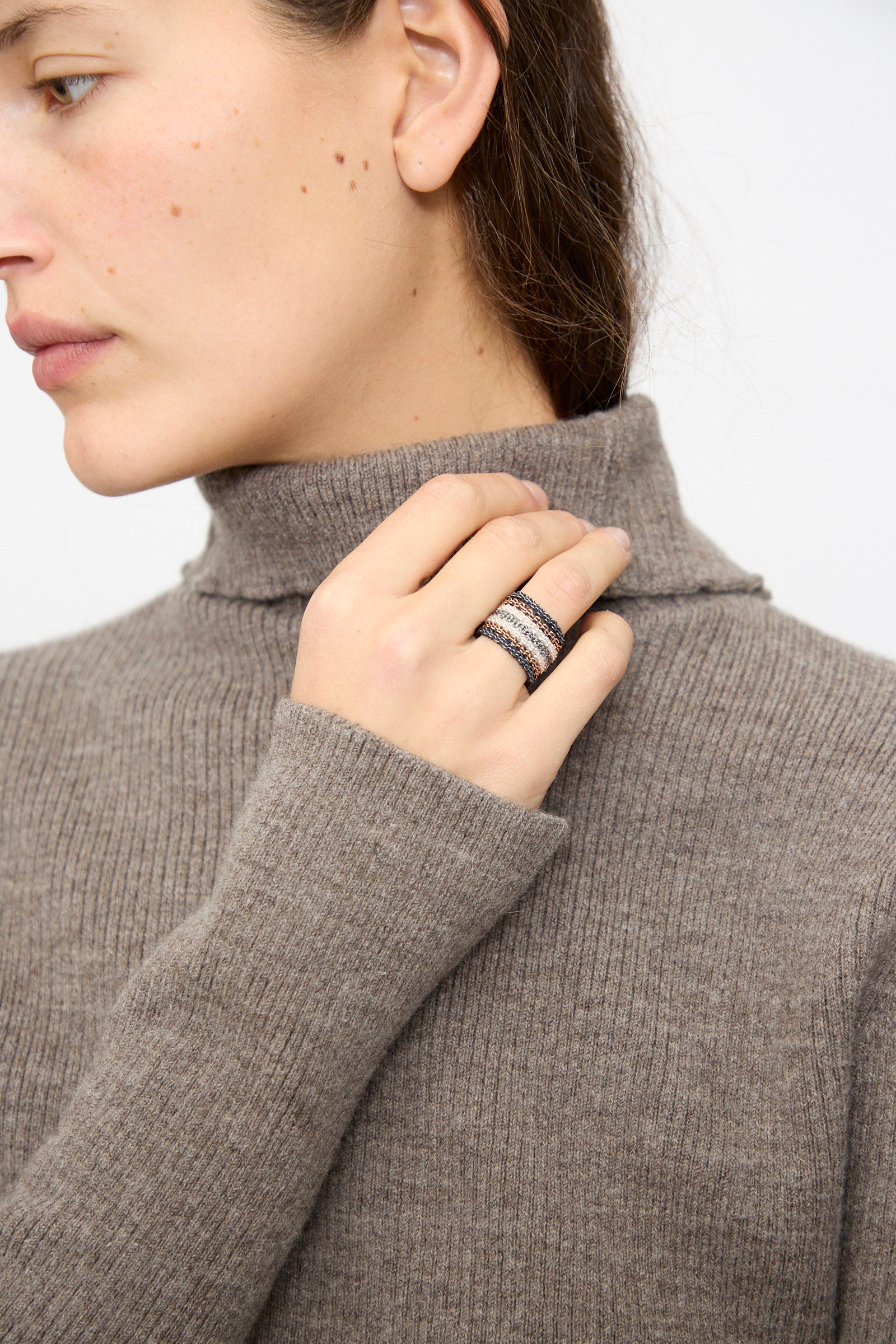 A person wears a taupe ribbed turtleneck sweater and raises their hand to display multiple stacked rings, featuring the striking Stephanie Schneider Silver, Rose Gold Plated and Silk Woven Ring.