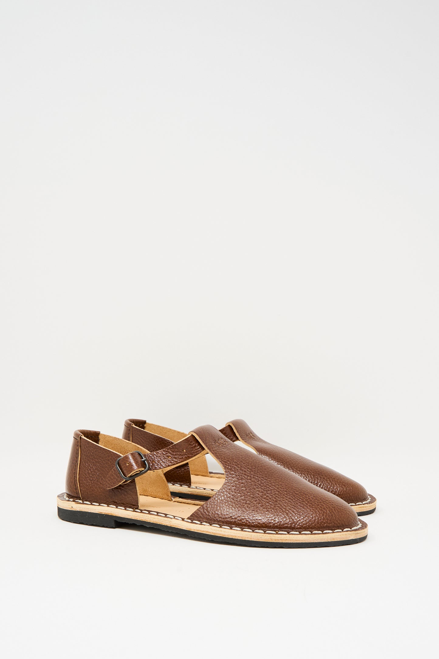 The Steve Mono Artisanal Sandal 10/12 in Brandy features vegetable-tanned leather, buckle straps, and flat recycled rubber soles, showcased on a plain white background.