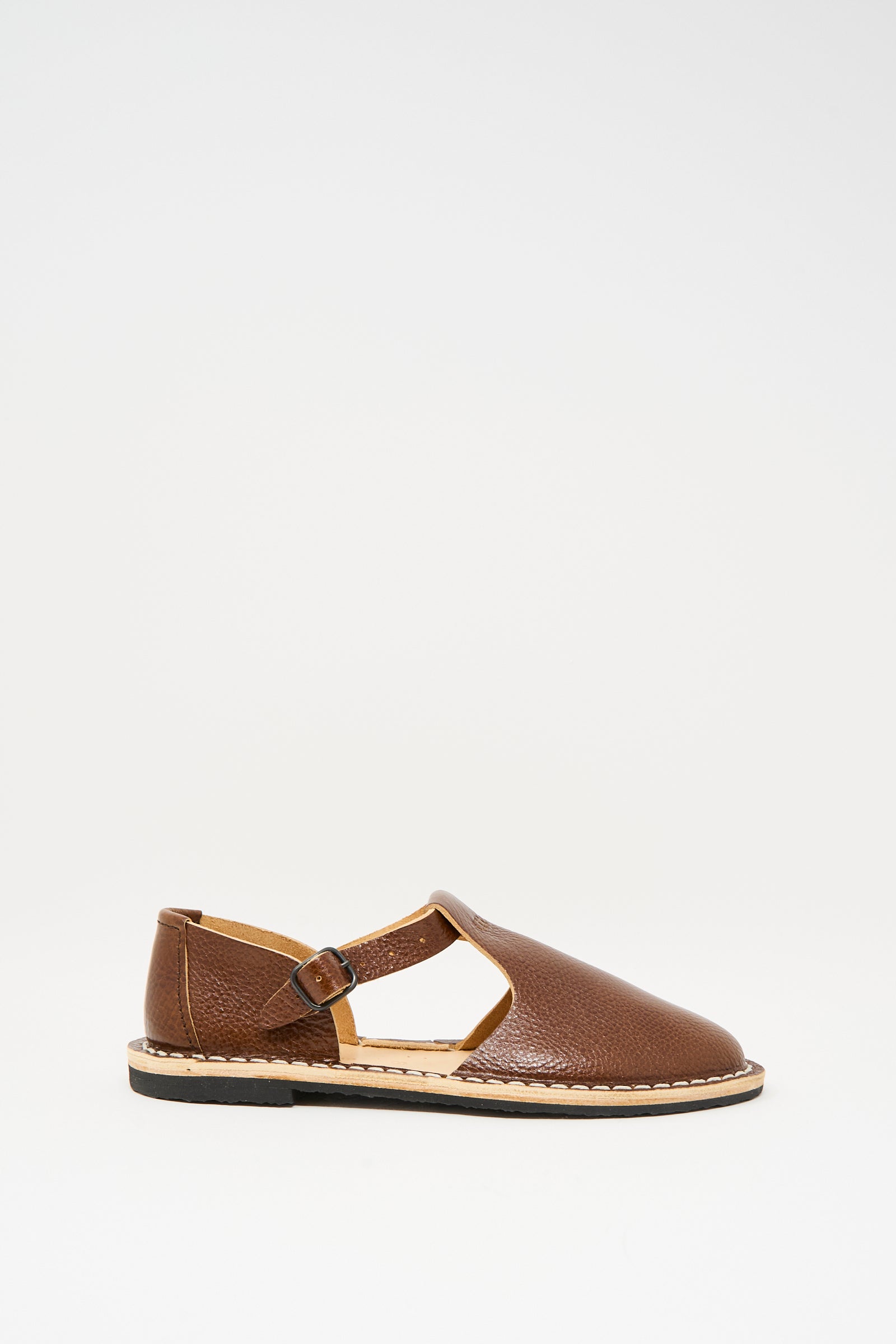 The Steve Mono Artisanal Sandal 10/12 in Brandy features brown vegetable-tanned leather, a closed toe, cut-out sides, side buckle strap, and a flat recycled rubber sole, shown against a white background.