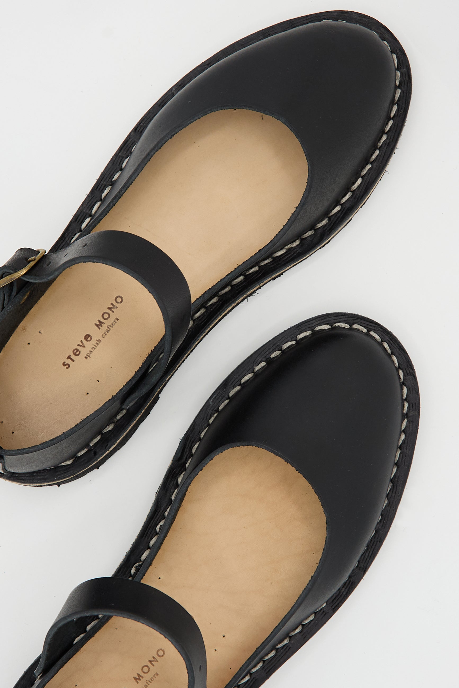 A pair of Vegetable Tanned Calfskin Artisanal Sandal in Black crafted from black vegetable tanned calfskin leather with visible stitching and a buckle strap. The insole is marked with the brand name "Steve Mono," highlighting its sustainably made origins.