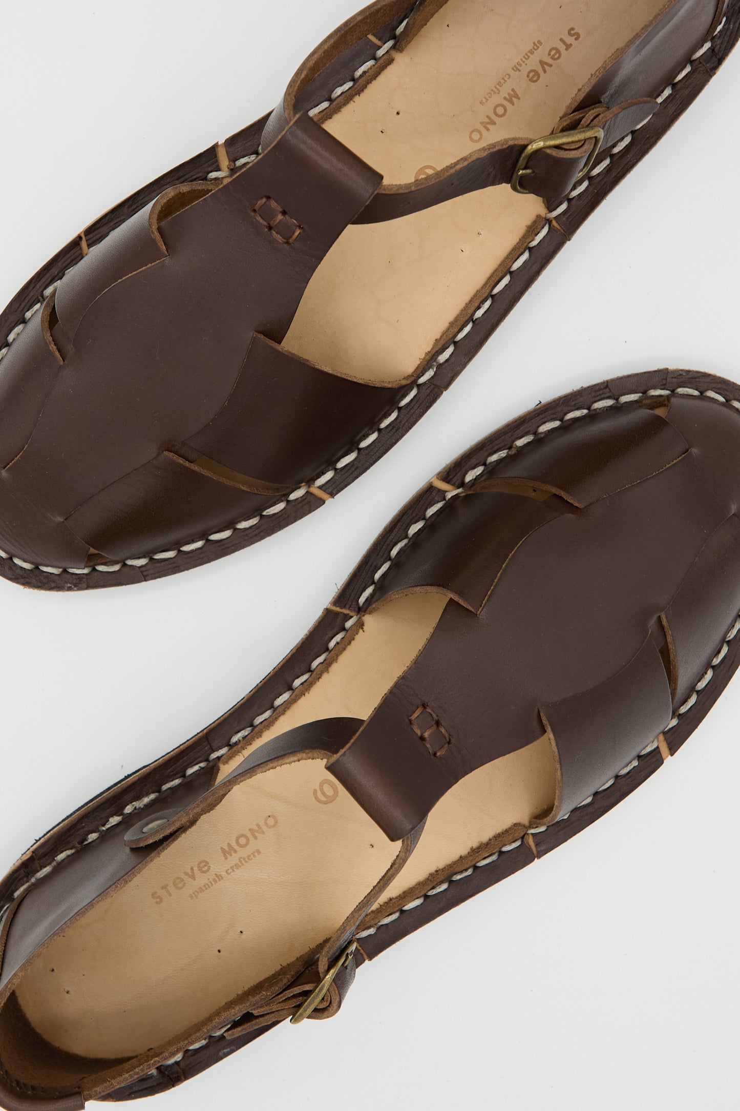 A pair of Steve Mono Vegetable Tanned Calfskin Artisanal Sandal in Coffee, featuring white stitching and buckle closures, displayed on a light background. These sustainably made sandals showcase a perfect blend of style and eco-conscious craftsmanship.