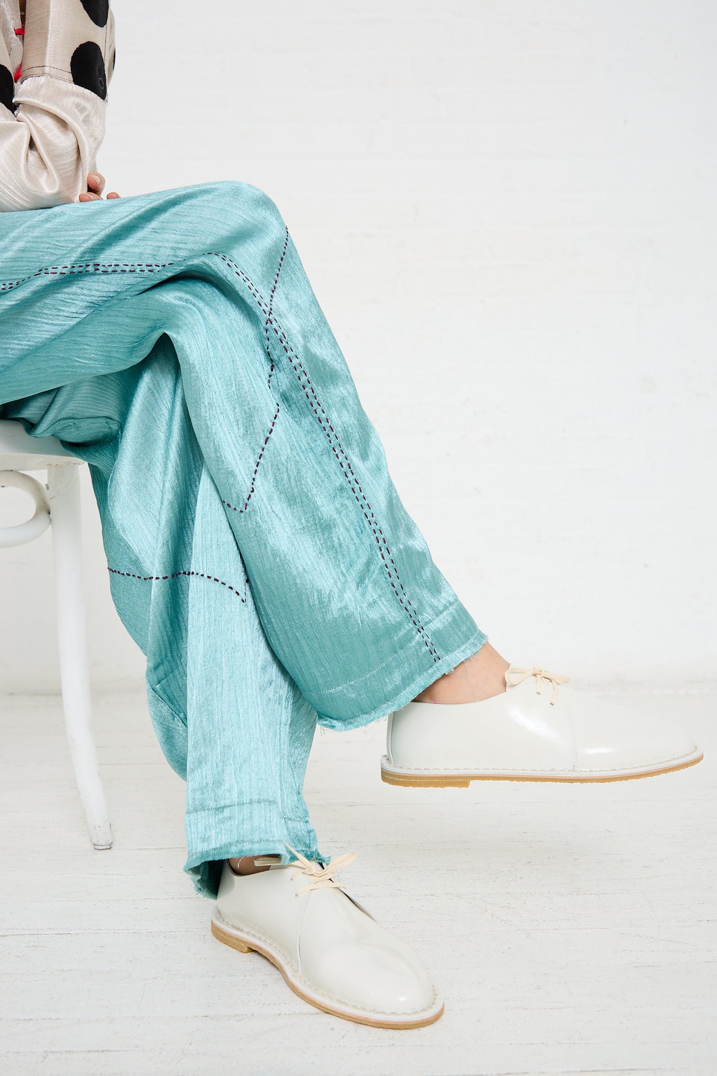 Person sitting on a white chair, wearing turquoise pants with a stitched pattern and sustainably made in Spain Patent Leather Derbie in White by Steve Mono featuring beige soles.
