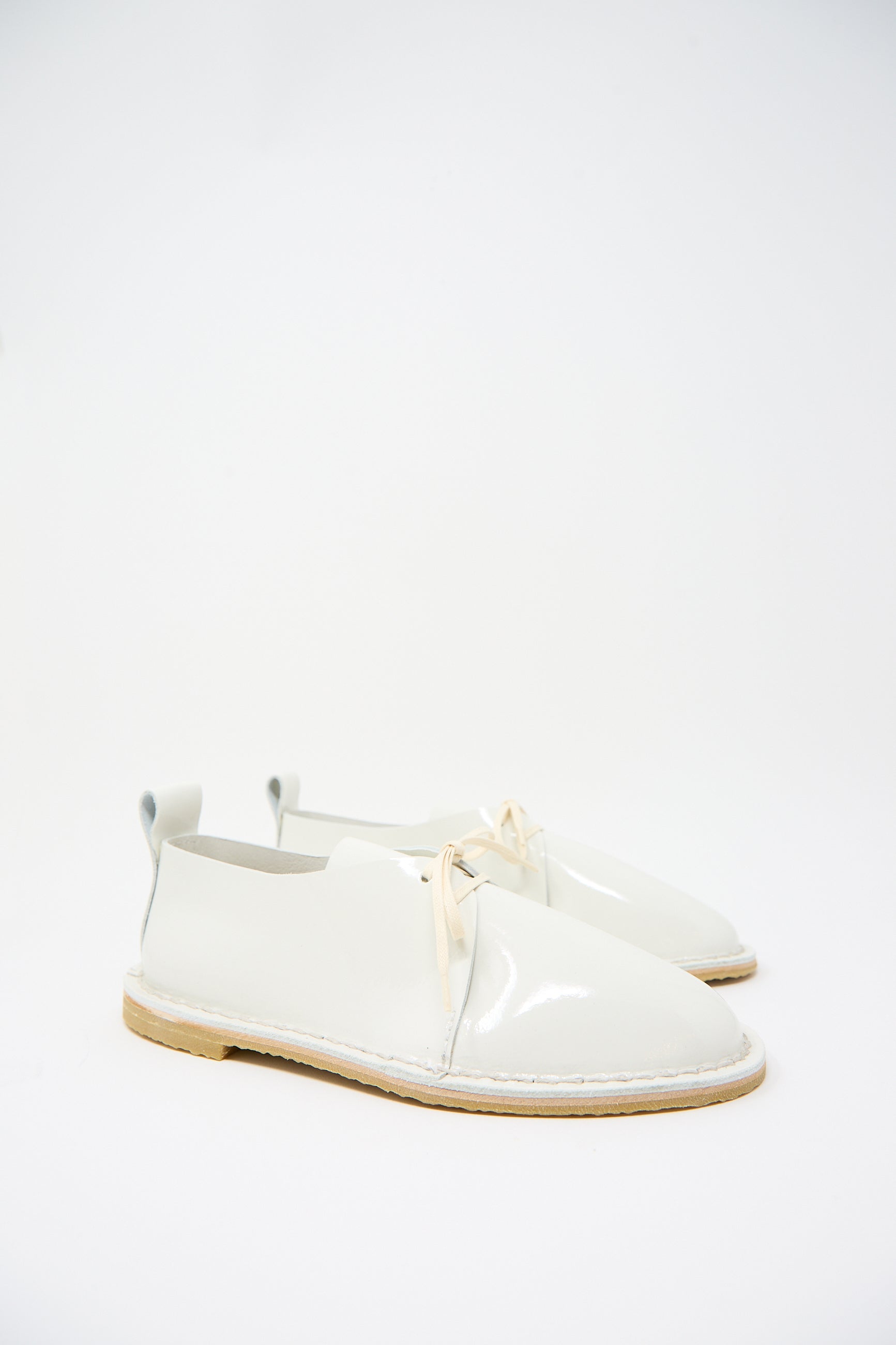 A pair of sustainably made white patent leather shoes with beige soles and laces are displayed on a plain white background. The Patent Leather Derbie in White by Steve Mono features a casual design with a pull tab at the back.