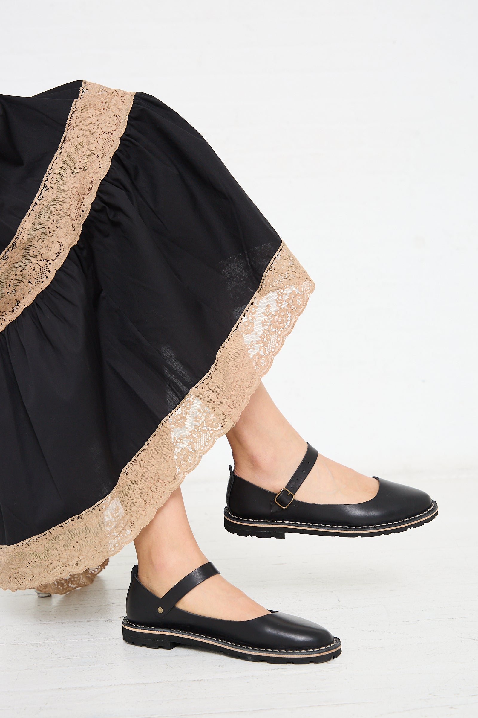 A person wearing a long black skirt with lace trim and black Steve Mono Vegetable Tanned Calfskin Artisanal Sandals in Black, crafted from sustainably made vegetable tanned leather, is standing on a white floor.