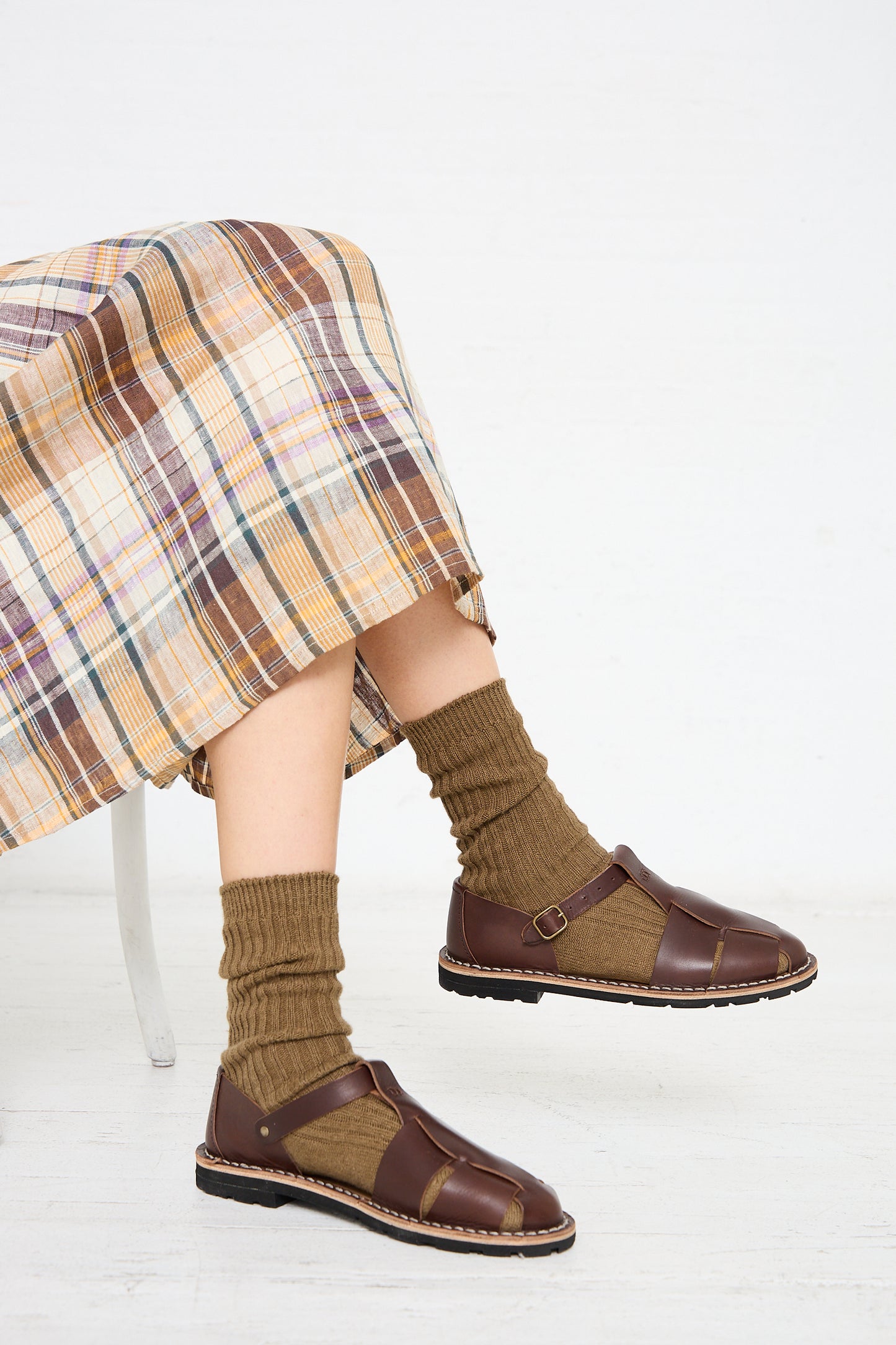 Person seated, wearing a plaid skirt, brown socks, and Steve Mono's Vegetable Tanned Calfskin Artisanal Sandal in Coffee. The white background gives a minimalistic setting.