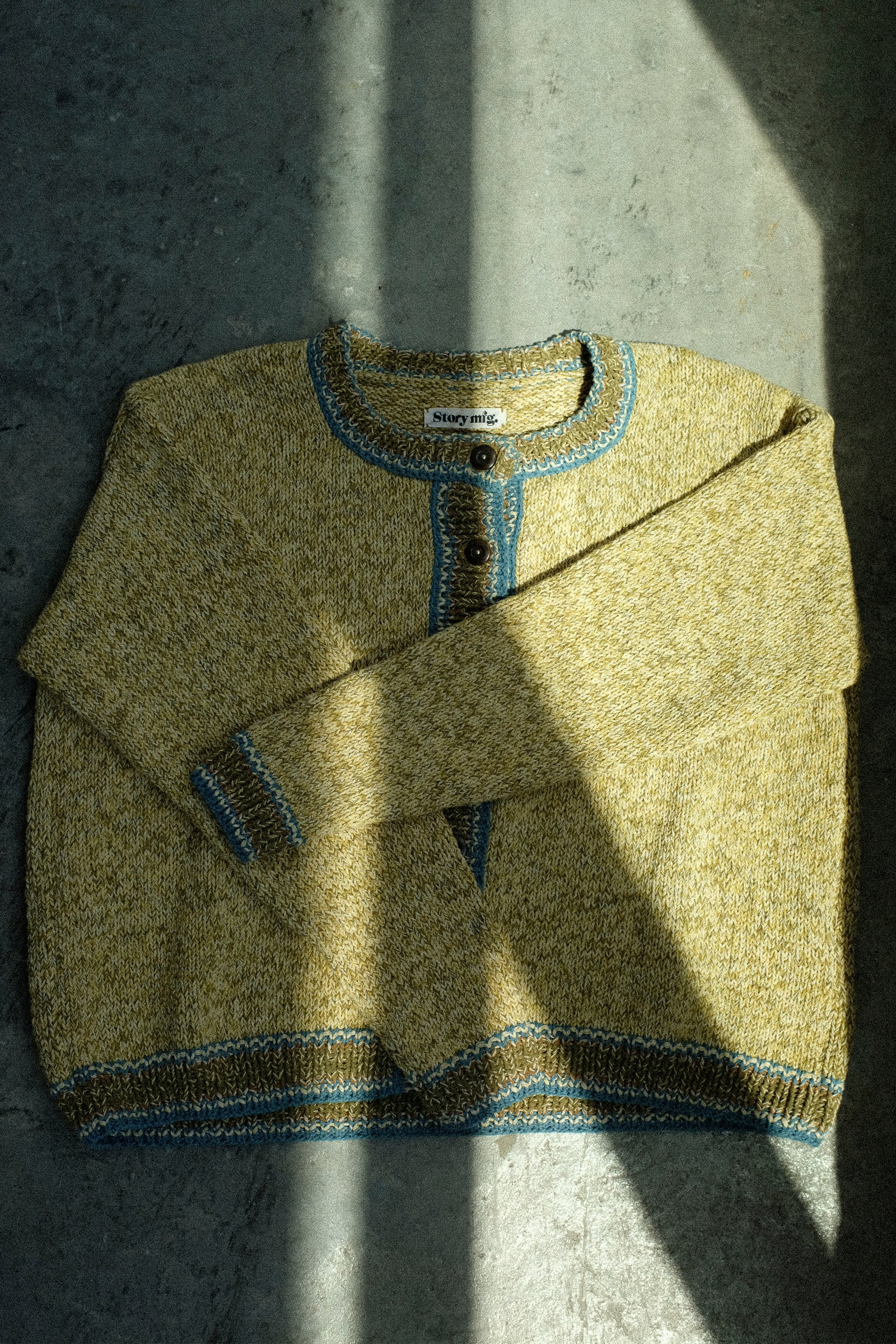 A flat image of Story mfg.'s Hand Knit Root Cardigan in Kelp on a concrete floor in sunlight with shade.