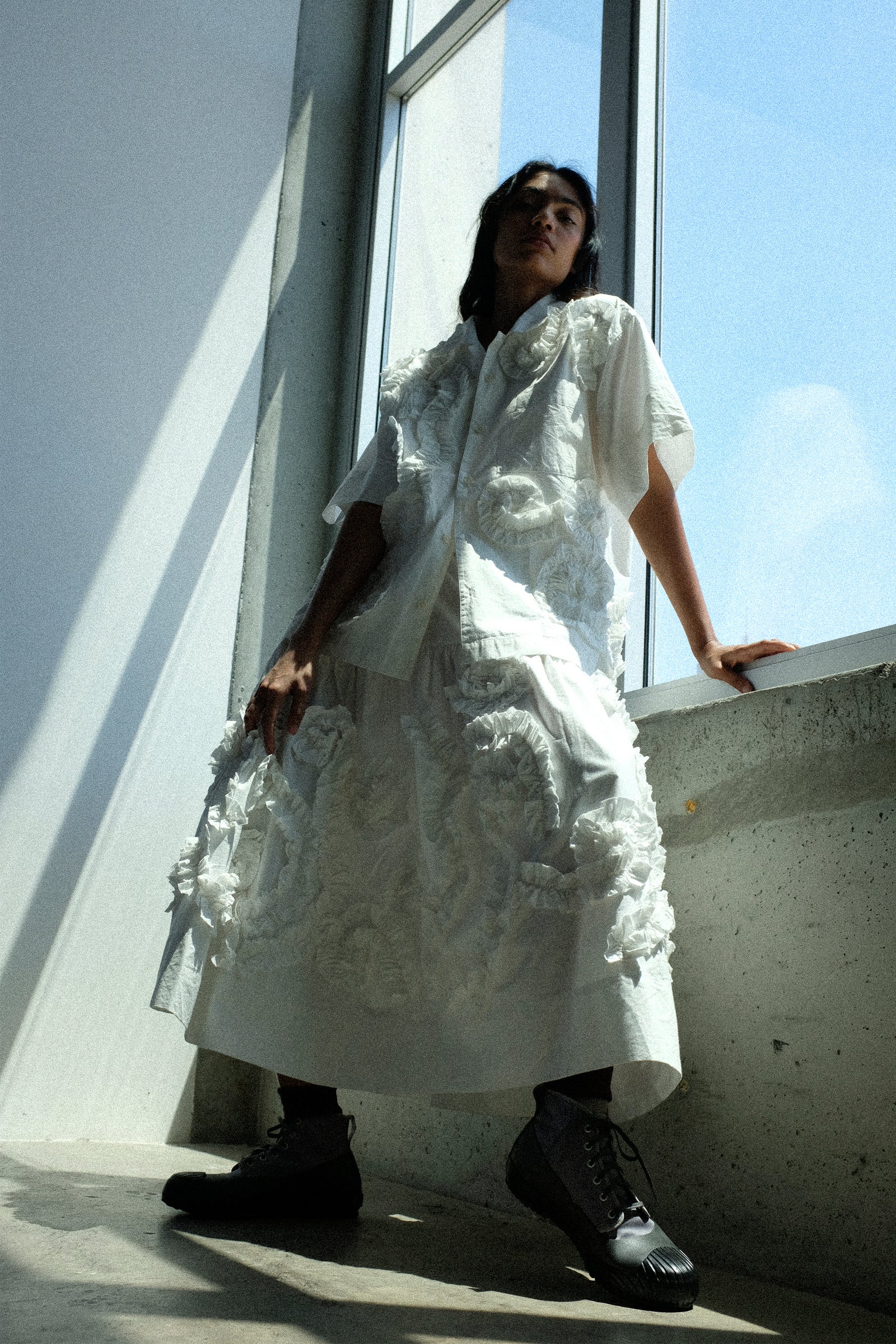 Person wearing Story mfg. Hand Applique Greetings Shirt in Ecru Fossil, with the Hand Applique Orbit Skirt in Ecru Fossil, standing against a modern building with large windows.