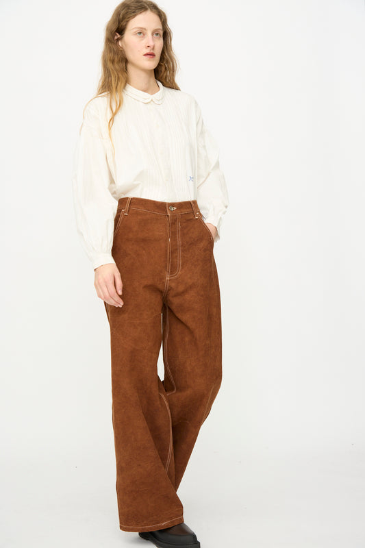 A person wears the Story mfg. Canvas Home Pant in Brown Wonky-Wear, paired with an oversized white collared shirt, standing against a plain white background.