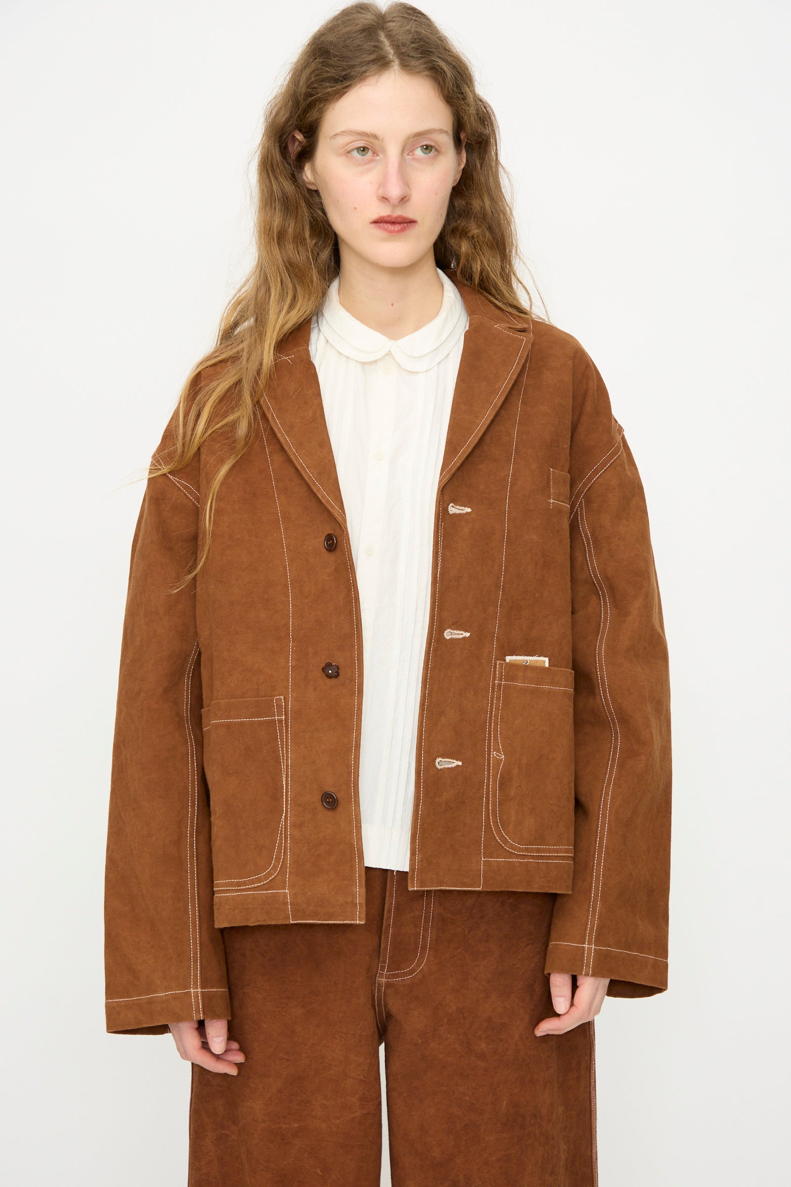 A person with long wavy hair wears the Story mfg. Canvas Cove Jacket in Brown Wonky-Wear over a white collared shirt and matching pants, standing against a plain light background.