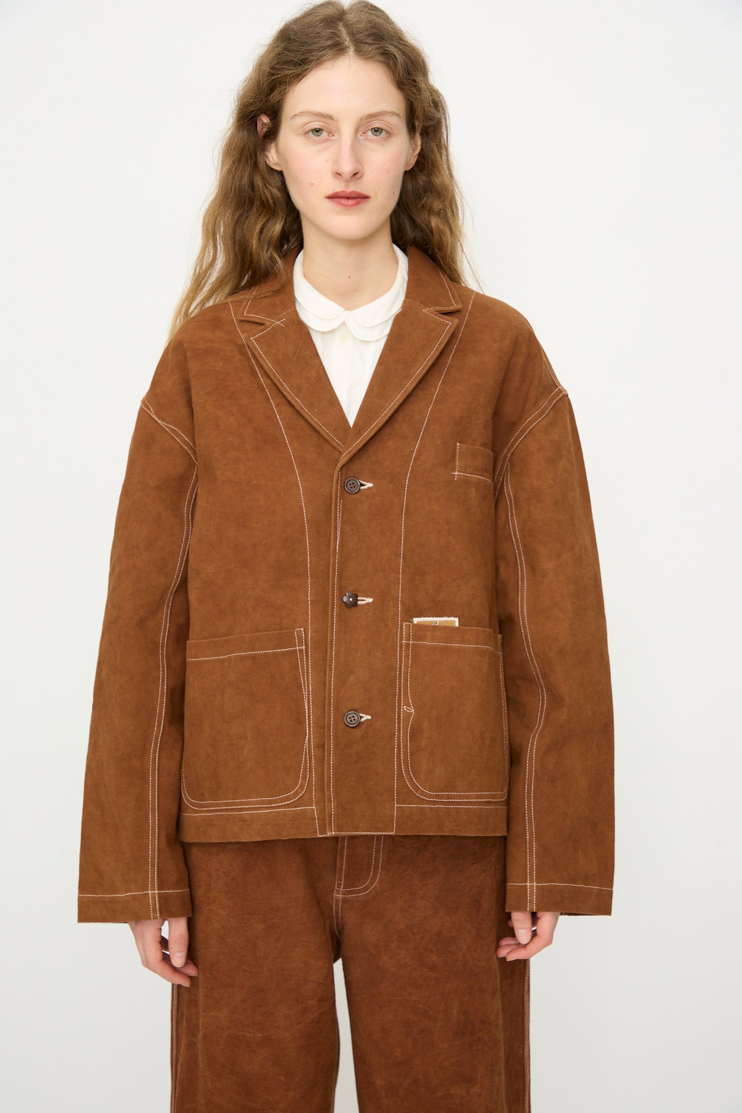Person with long wavy hair wears the Story mfg. Canvas Cove Jacket in Brown Wonky-Wear, featuring white stitching, standing against a plain white background.