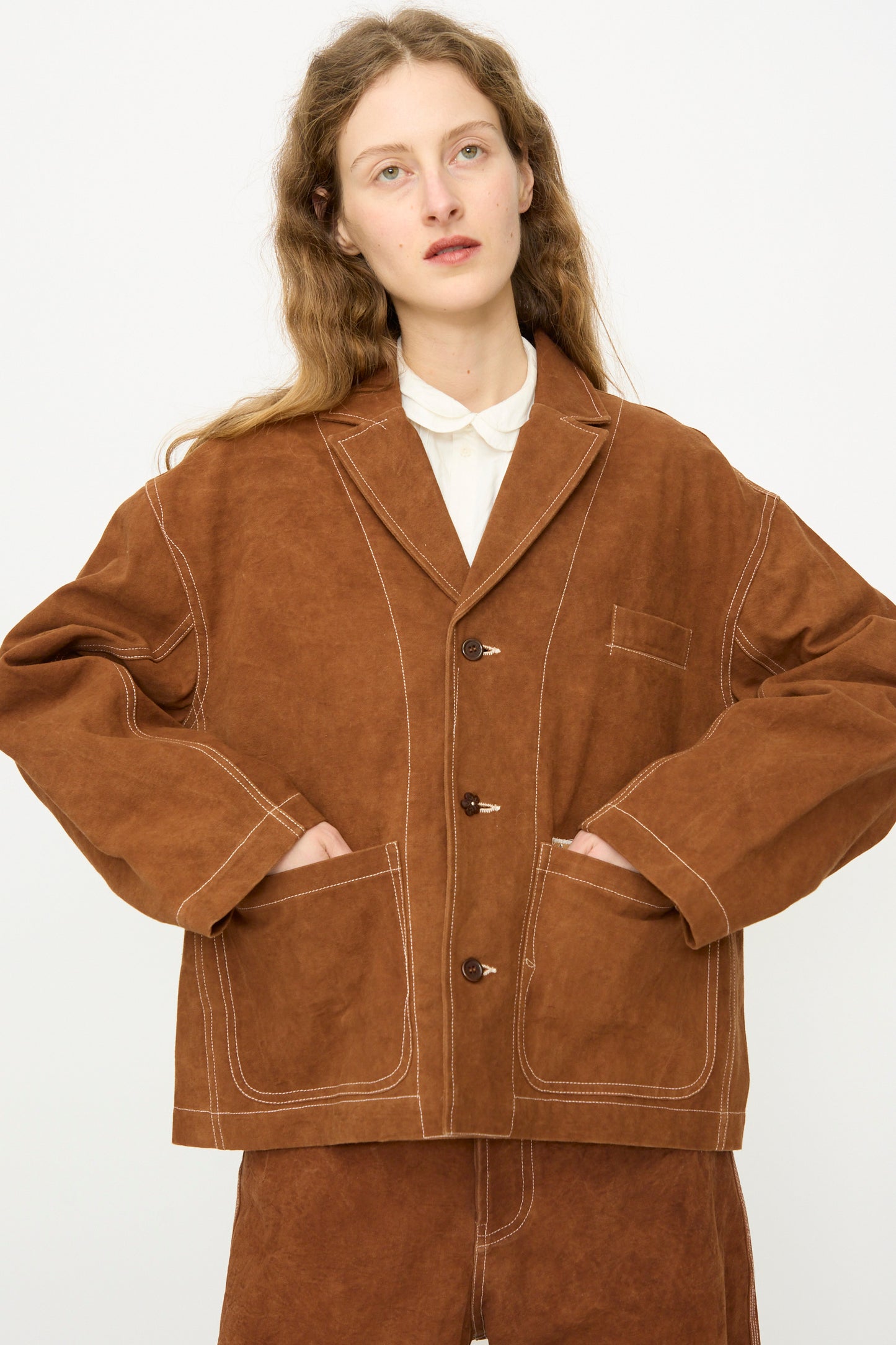 A person with long wavy hair models the Story mfg. Canvas Cove Jacket in Brown Wonky-Wear, featuring large front pockets, paired with matching pants, against a plain white background.