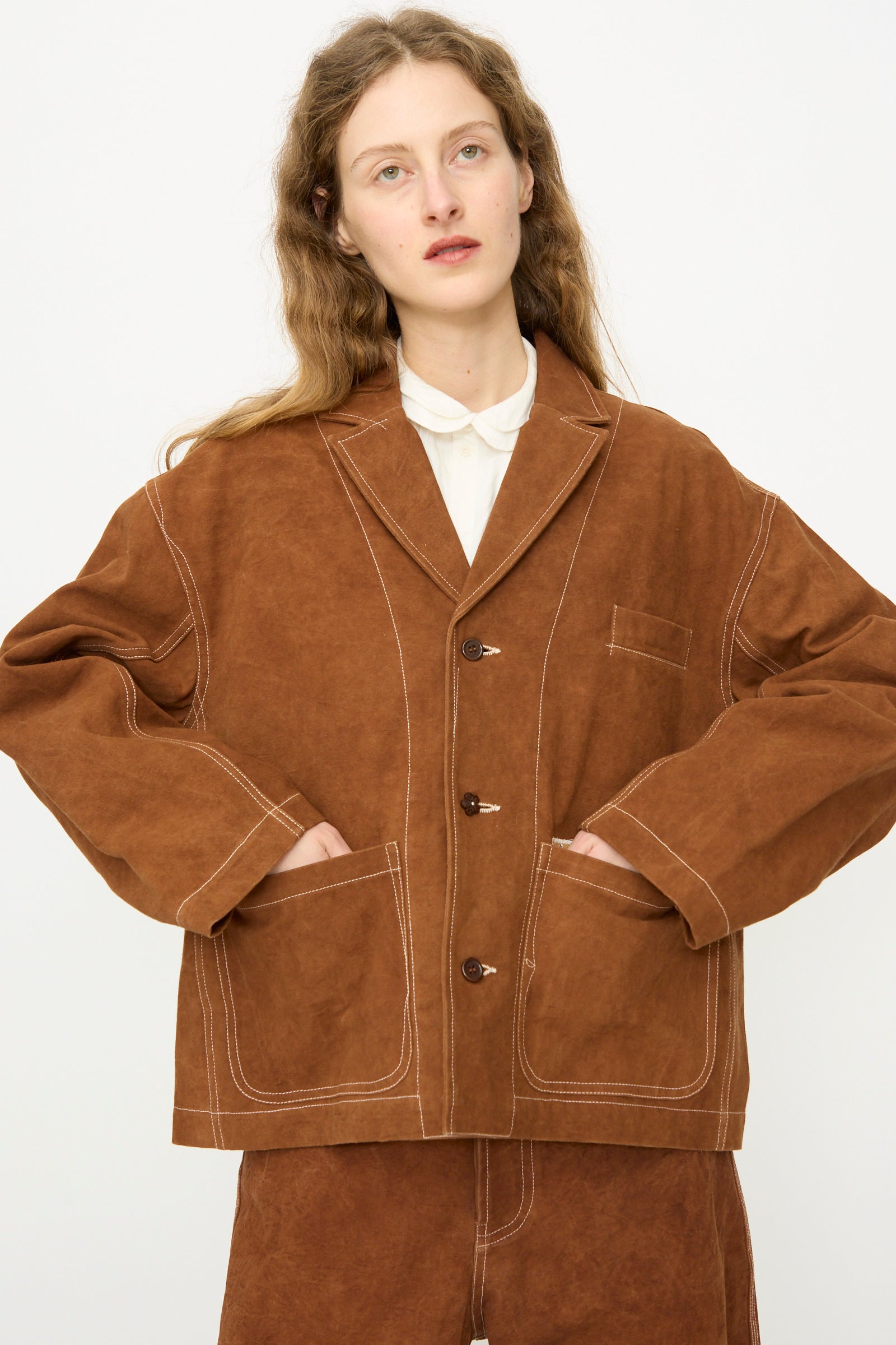 A person with long wavy hair models the Story mfg. Canvas Cove Jacket in Brown Wonky-Wear, featuring large front pockets, paired with matching pants, against a plain white background.