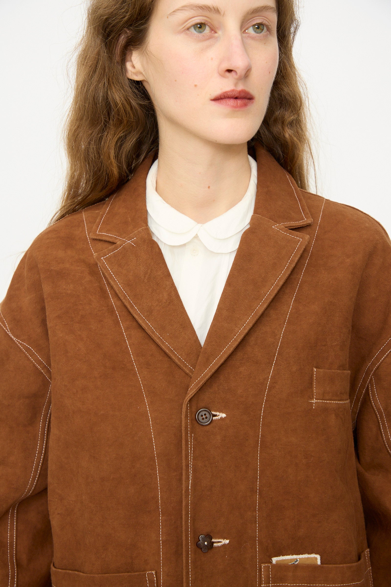 Someone wears the Story mfg. Canvas Cove Jacket in Brown Wonky-Wear, featuring white stitching over a white collared shirt, photographed from the chest up against a plain background.