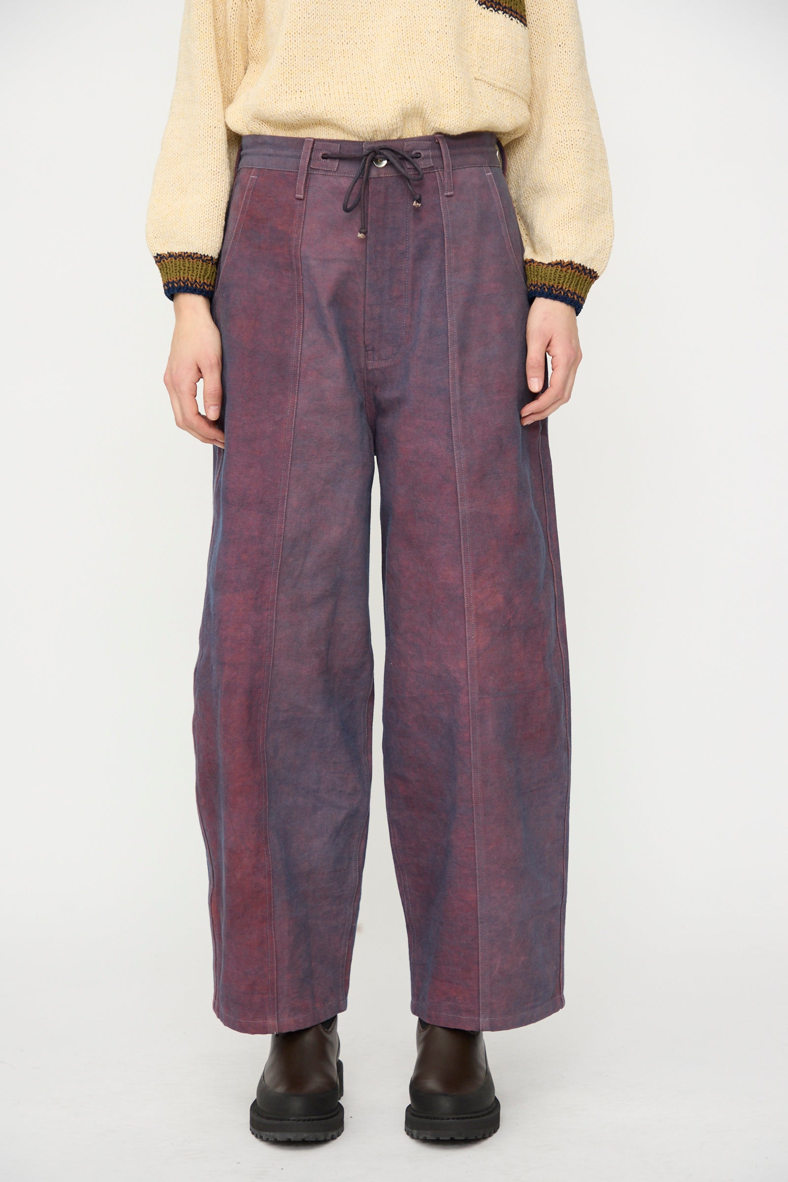 A person stands against a plain background wearing a beige sweater, black boots, and Story mfg.’s Canvas Document Pant in Ancient Purple.