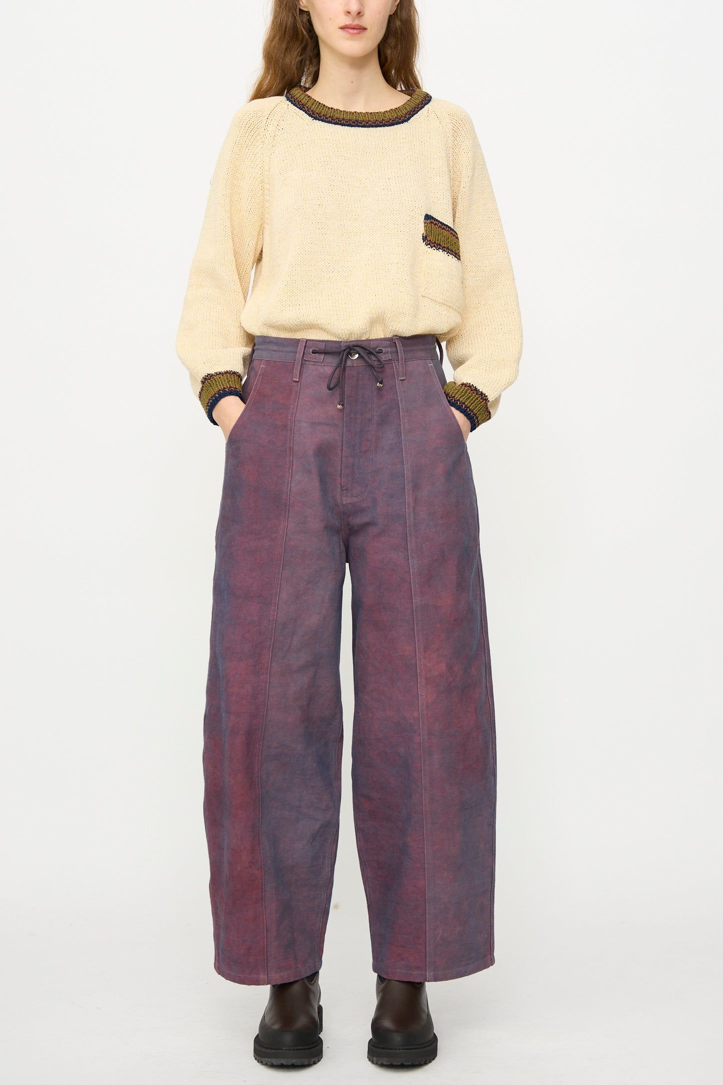 A person stands against a plain white background wearing a beige sweater with pocket detail, Story mfg.'s Canvas Document Pant in Ancient Purple—relaxed fit, wide-leg with drawstring waist—and dark ankle boots.