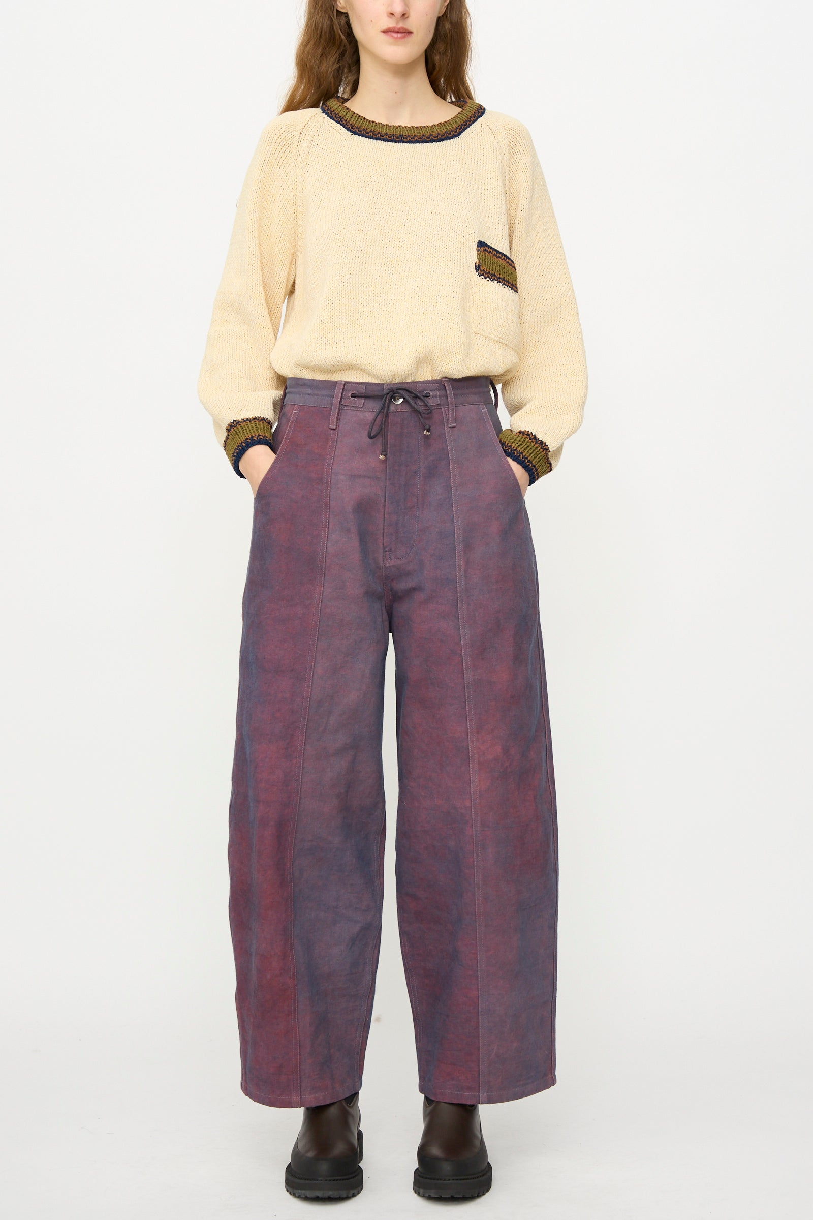 A person stands against a plain white background wearing a beige sweater with pocket detail, Story mfg.'s Canvas Document Pant in Ancient Purple—relaxed fit, wide-leg with drawstring waist—and dark ankle boots.