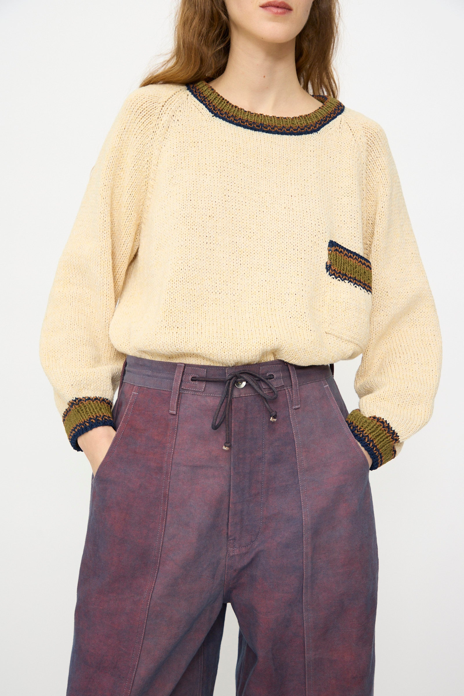 A person wears a cream knit sweater with colored trim and a small chest pocket, paired with Story mfg.'s Canvas Document Pant in Ancient Purple, hands in pockets.
