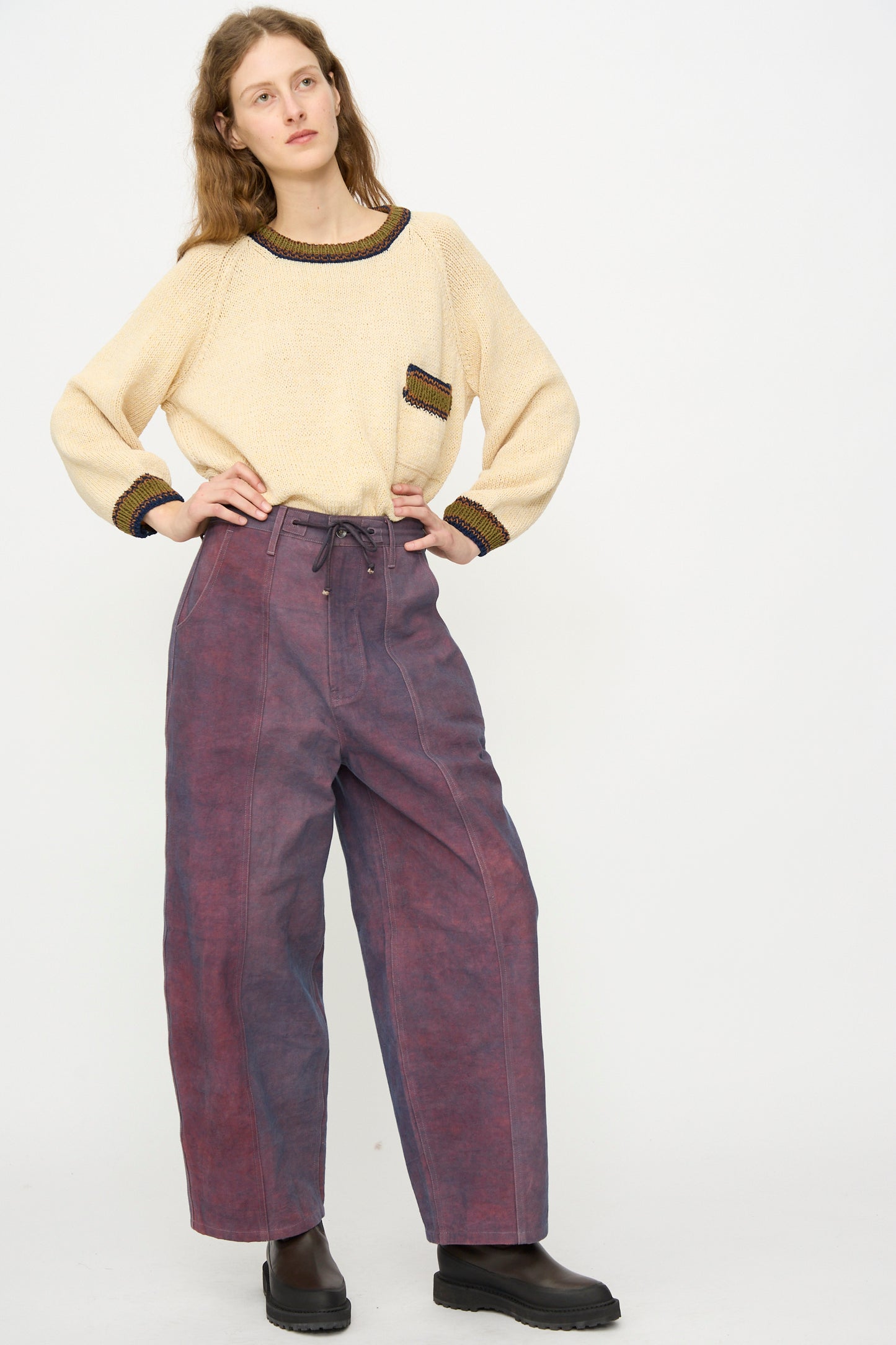 A woman stands against a plain background wearing a beige sweater with dark trim, Story mfg.'s Canvas Document Pant in Ancient Purple, and black shoes.