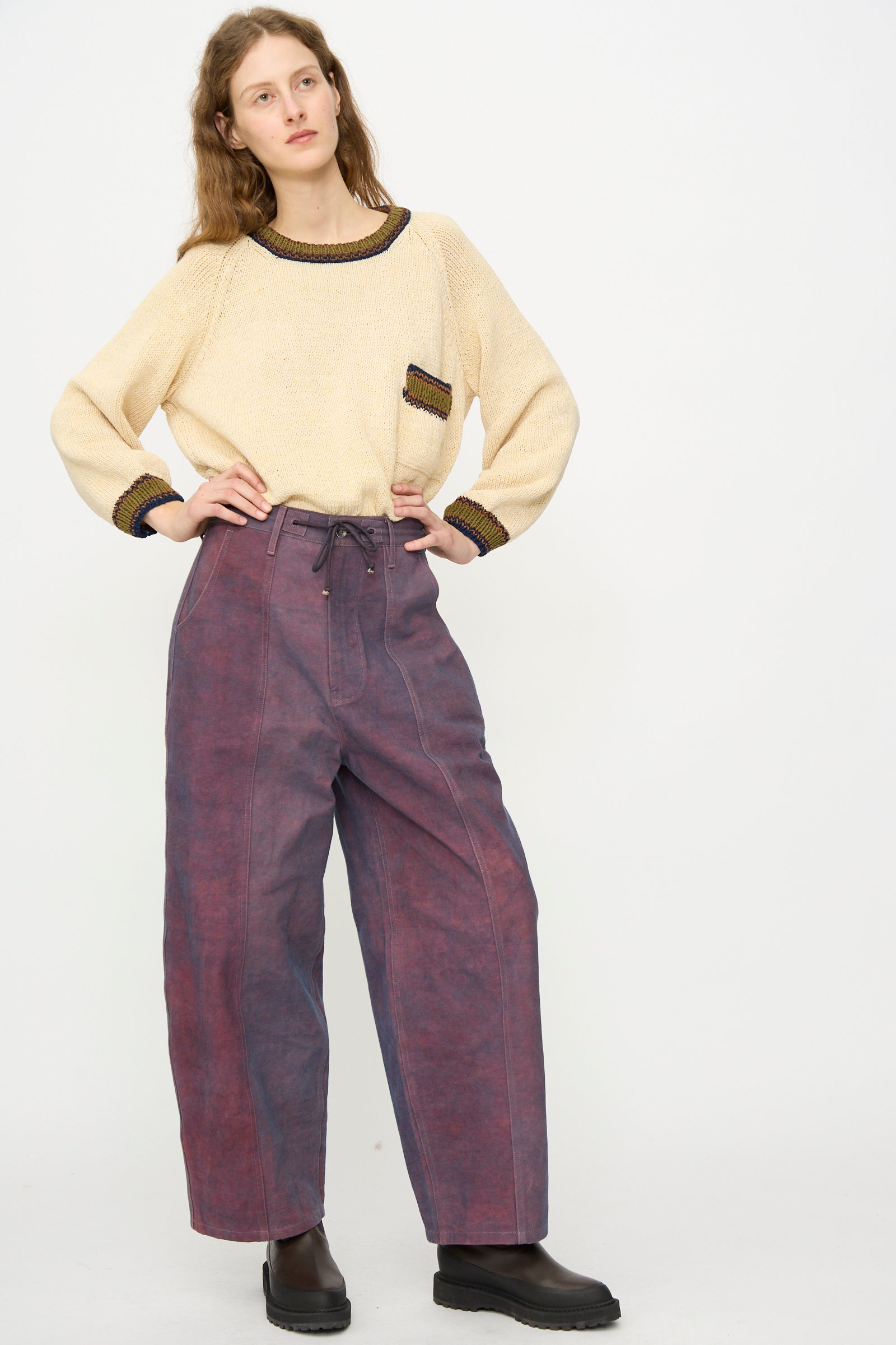 A woman stands against a plain background wearing a beige sweater with dark trim, Story mfg.'s Canvas Document Pant in Ancient Purple, and black shoes.