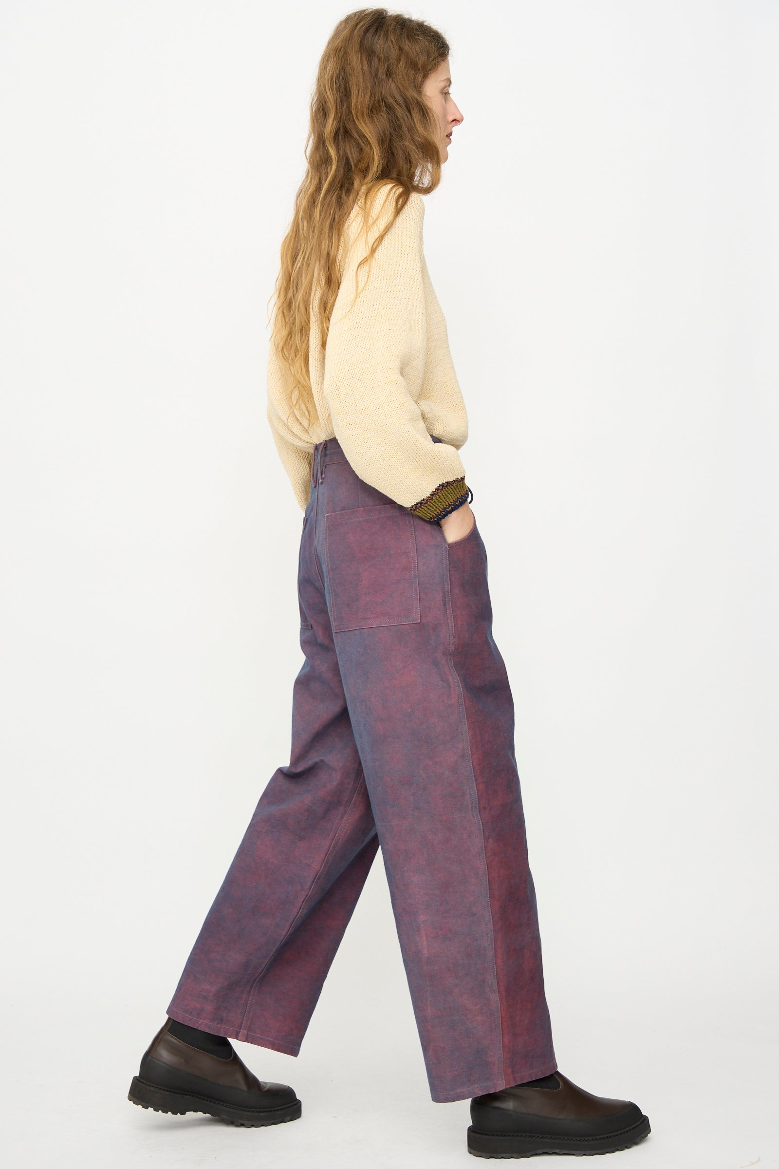 A person wearing Story mfg.'s Canvas Document Pant in Ancient Purple and a beige sweater stands in profile with hands in pockets against a plain white background.