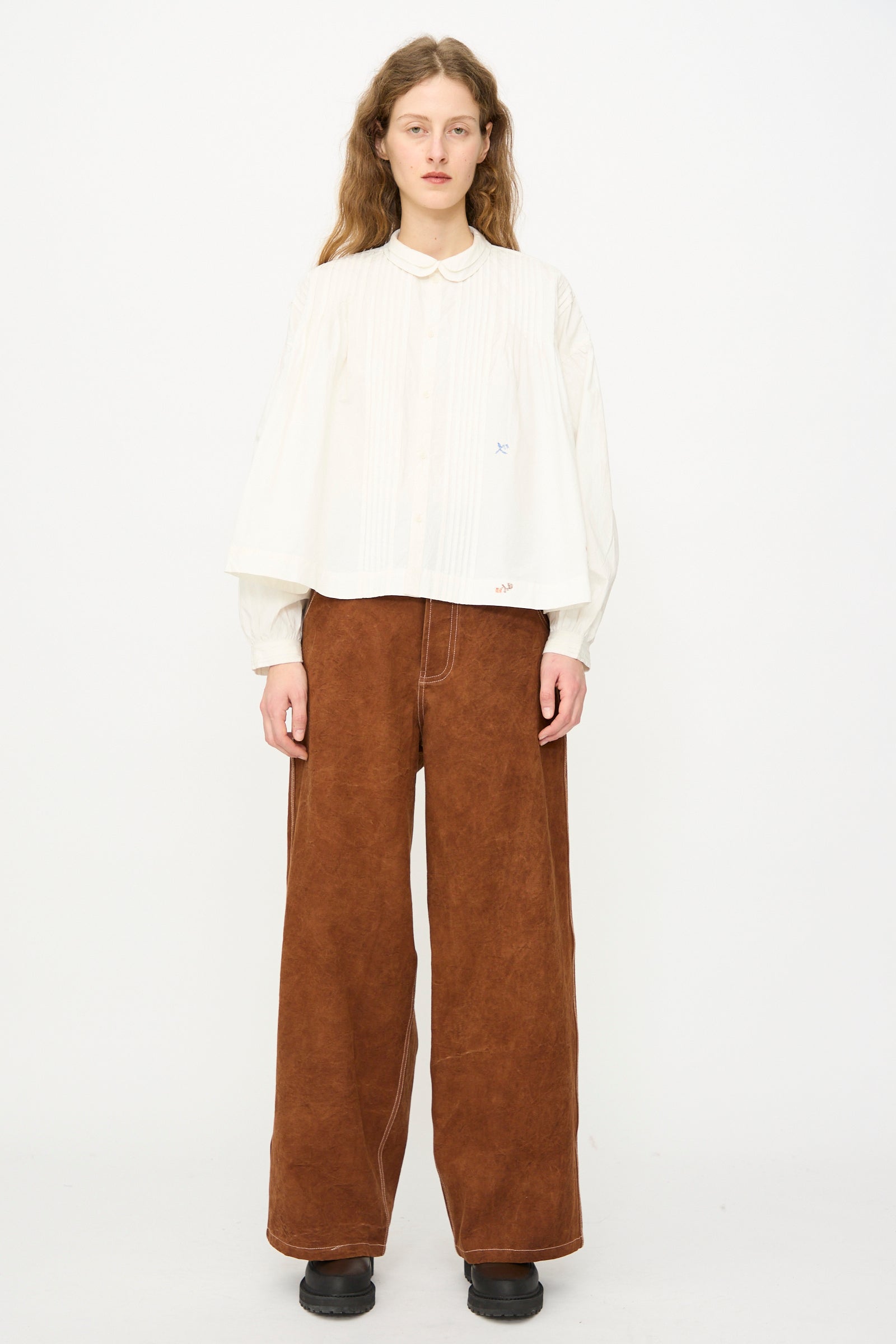A person stands against a plain background, wearing an oversized white organic cotton blouse, the Story mfg. Canvas Home Pant in Brown Wonky-Wear, and black shoes.