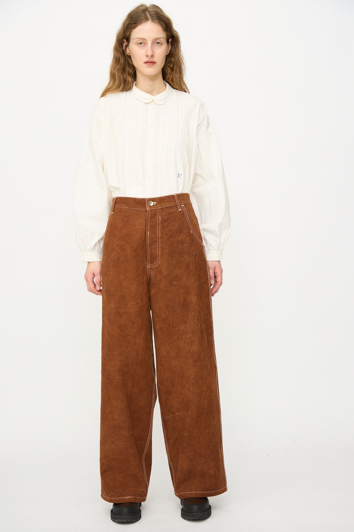 A person stands against a plain background wearing an oversized white long-sleeve button-up shirt, black shoes, and Story mfg.'s Canvas Home Pant in Brown Wonky-Wear with white stitching.