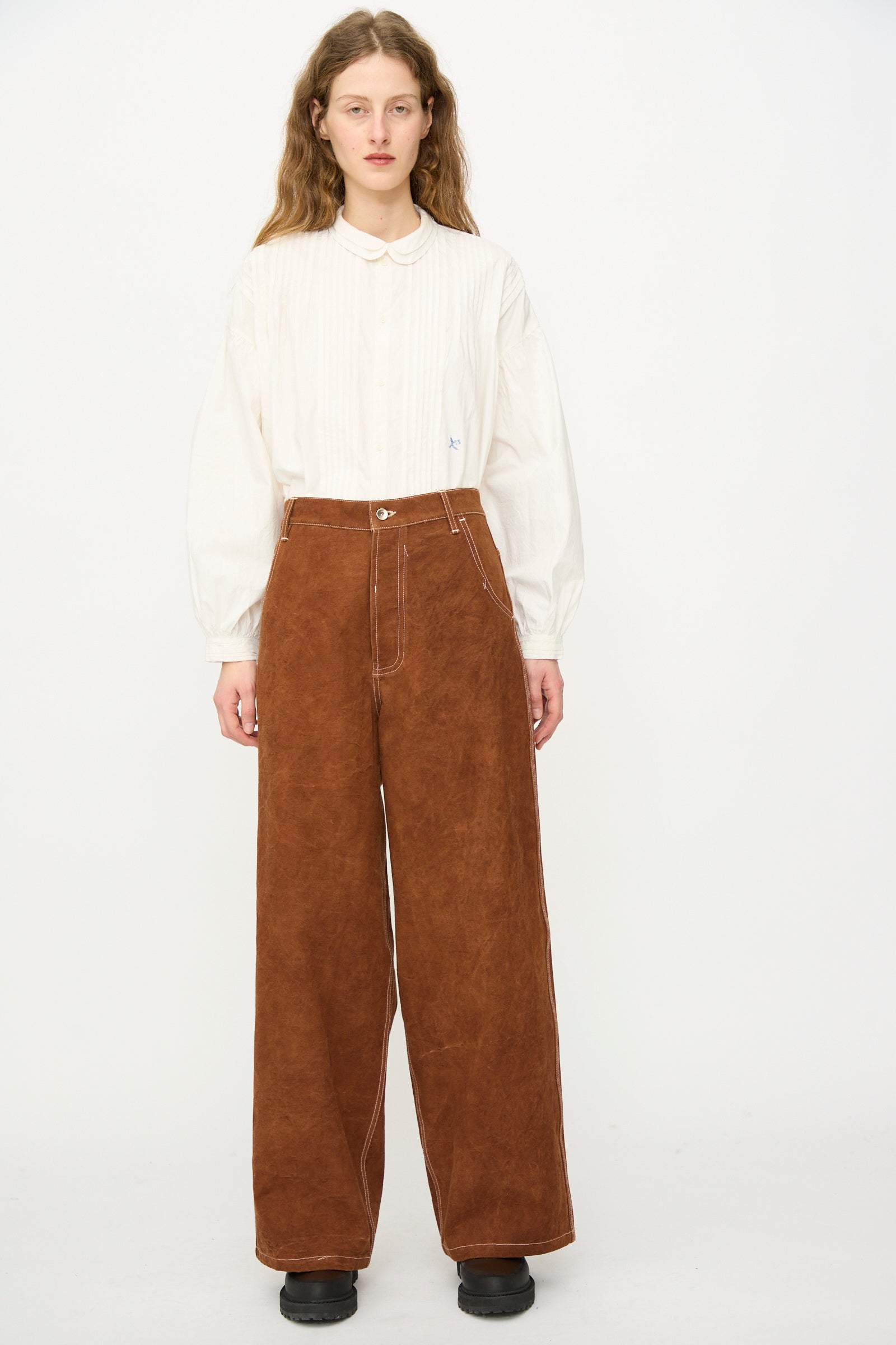 A person stands against a plain background wearing an oversized white long-sleeve button-up shirt, black shoes, and Story mfg.'s Canvas Home Pant in Brown Wonky-Wear with white stitching.