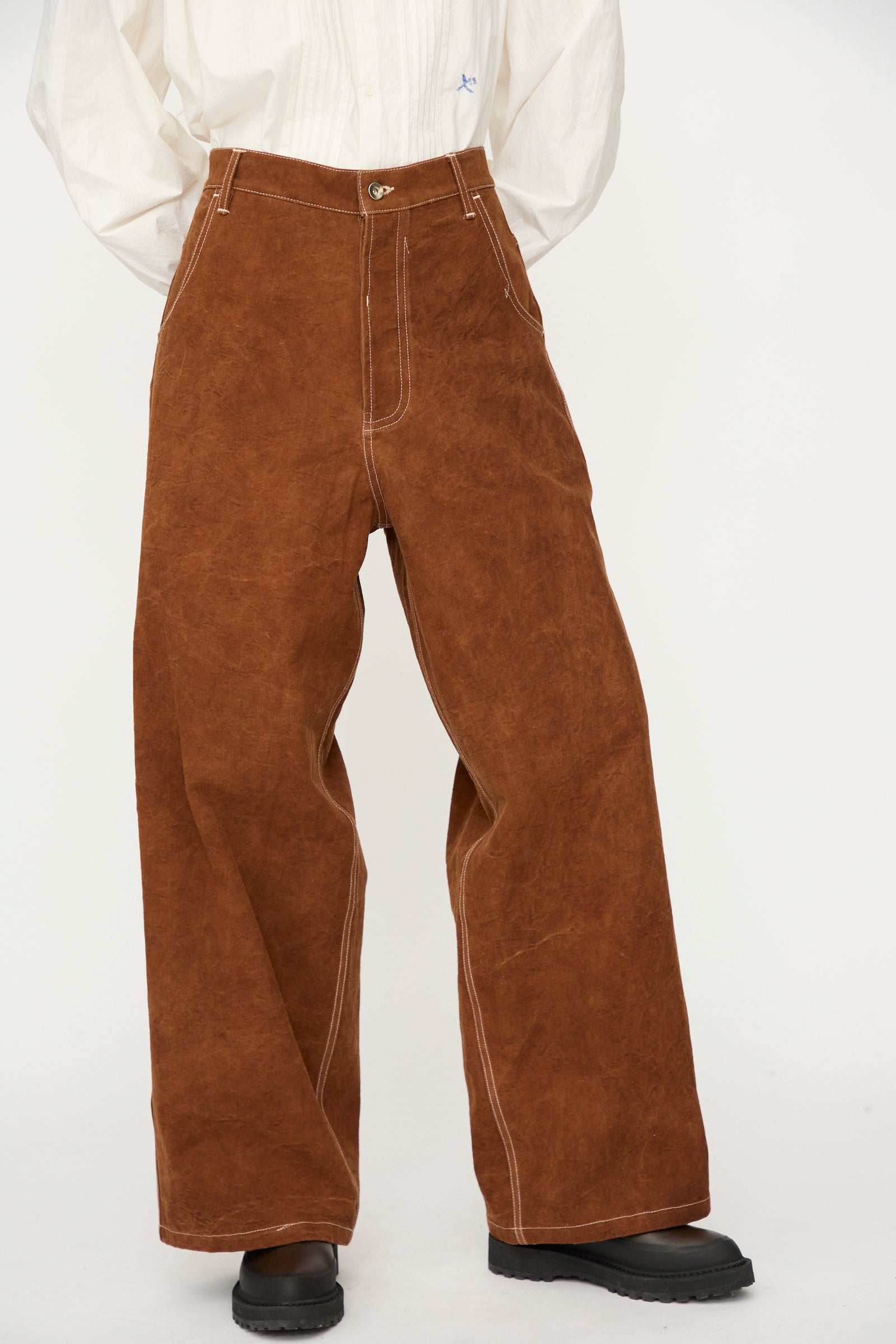 A person wears the Story mfg. Canvas Home Pant in Brown Wonky-Wear—oversized, wide-leg trousers with white stitching—paired with a tucked-in white shirt and black shoes, standing against a plain white background.